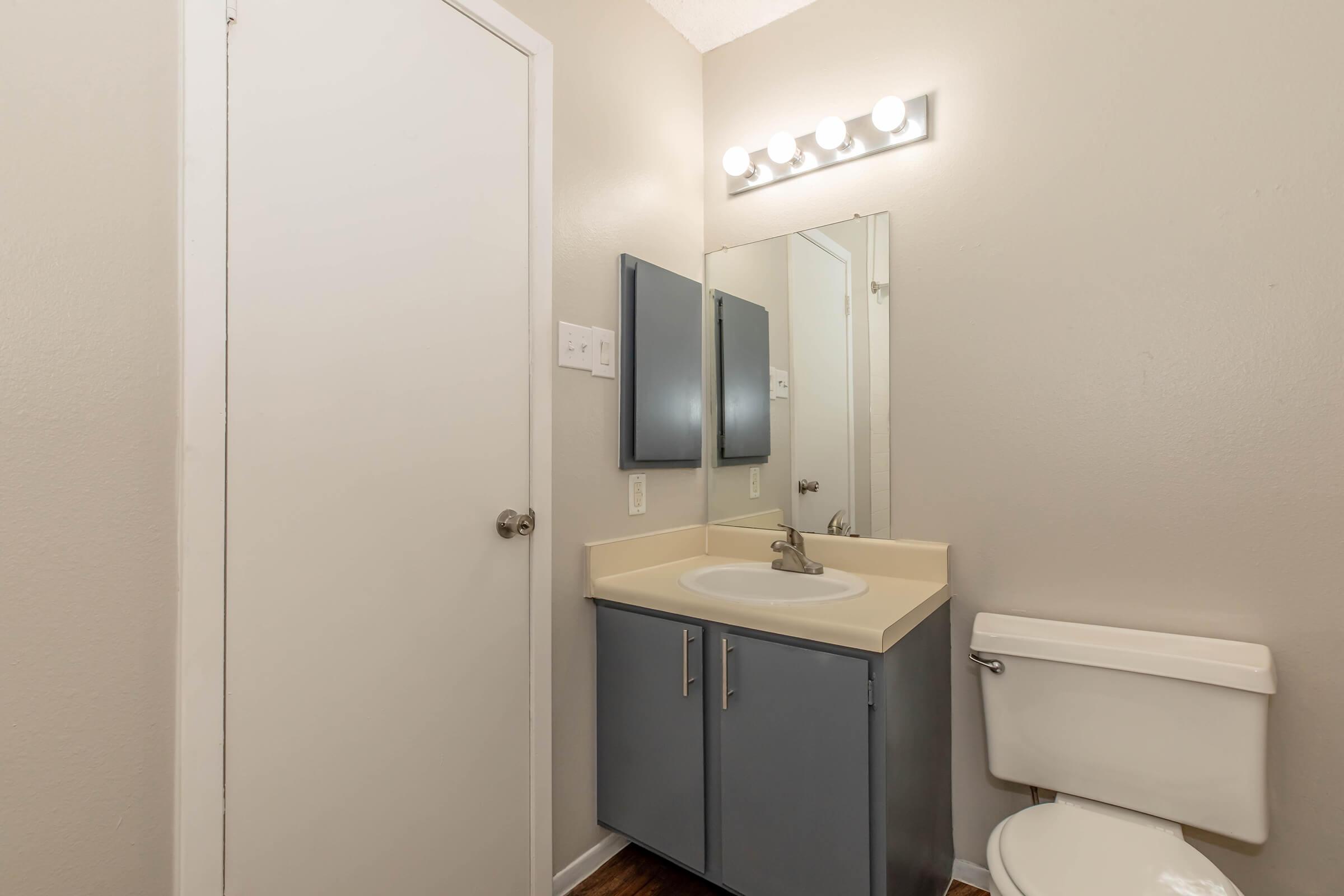 A simple bathroom featuring a light-colored wall, a plain mirror above a sink with a gray cabinet, a toilet, and a door. The lighting is provided by a row of light bulbs above the mirror, creating a bright and clean environment.