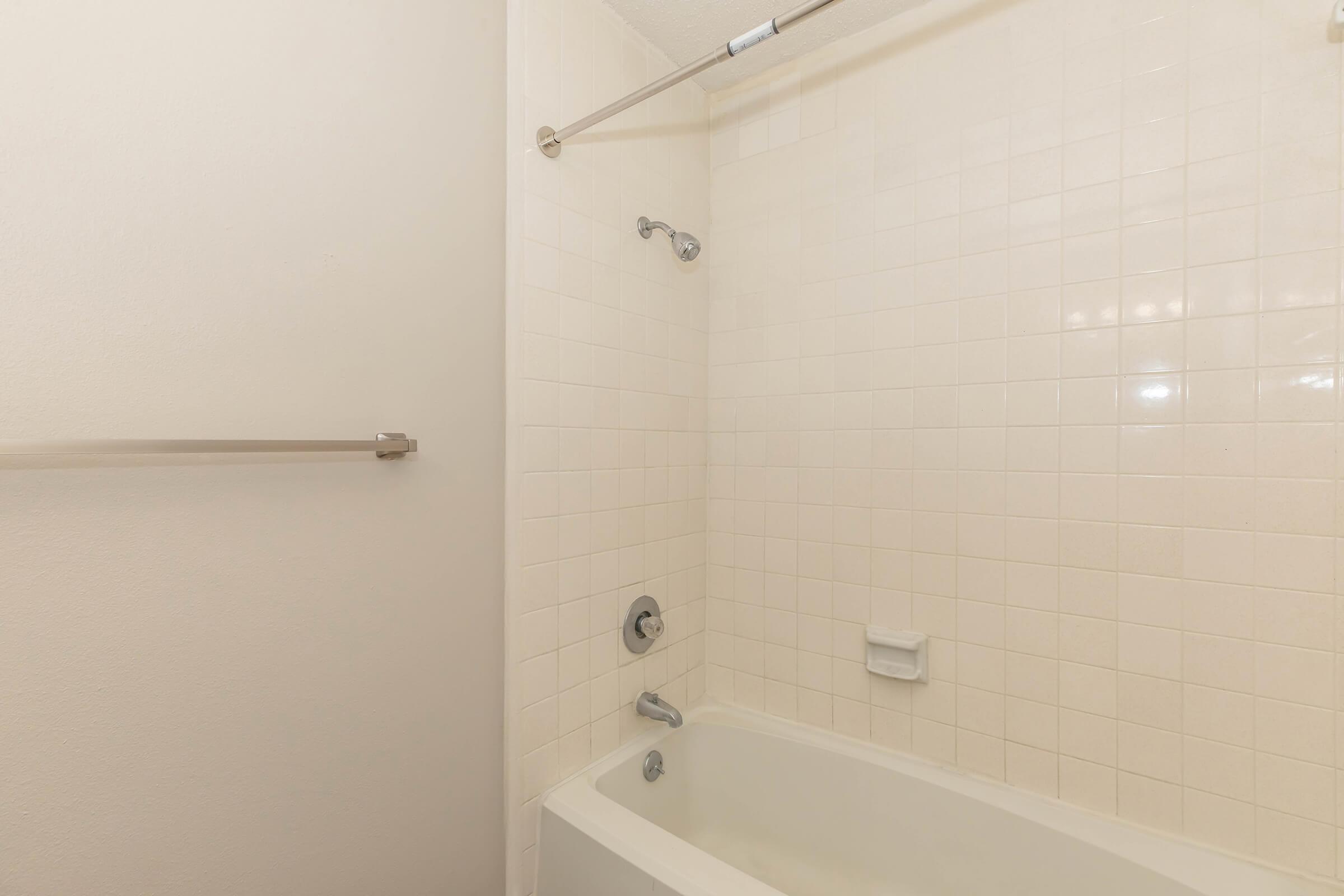A clean, minimalist bathroom featuring a white bathtub, a showerhead mounted on a wall, and a towel rack. The walls are tiled with light-colored square tiles, contributing to a simple and bright aesthetic. The lighting is soft, enhancing the serene atmosphere of the space.