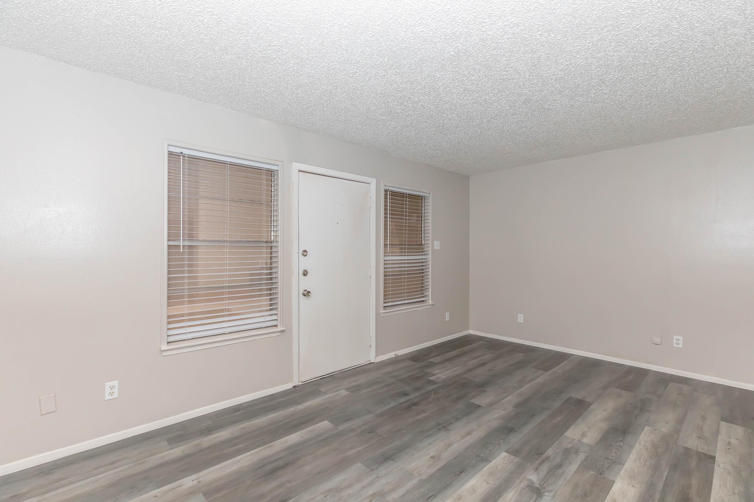 A spacious, empty room with light gray walls and textured ceiling. It features a front door with a window beside it on one wall and two smaller windows on the adjacent wall, allowing natural light. The floor is made of light-colored laminate wood, creating a clean and modern atmosphere.