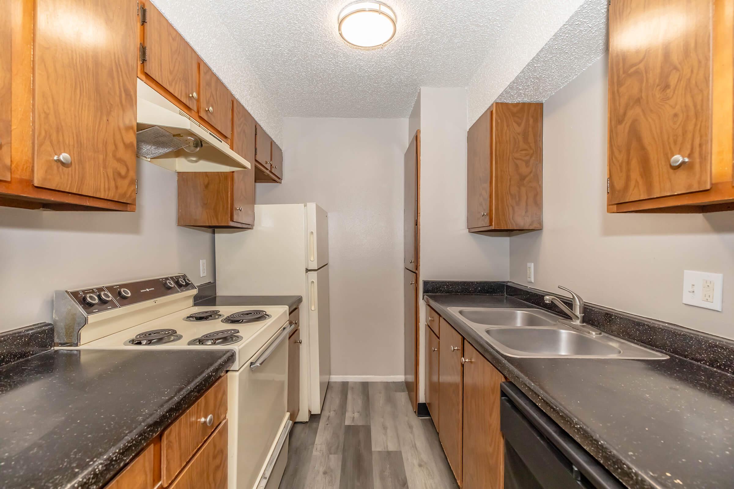 A view of a modern kitchen featuring wooden cabinetry, a white refrigerator, and a black countertop. The kitchen includes a stove with four burners, a double sink, and a light fixture on the ceiling. The floor is a light-colored laminate, enhancing the space's clean and contemporary look.