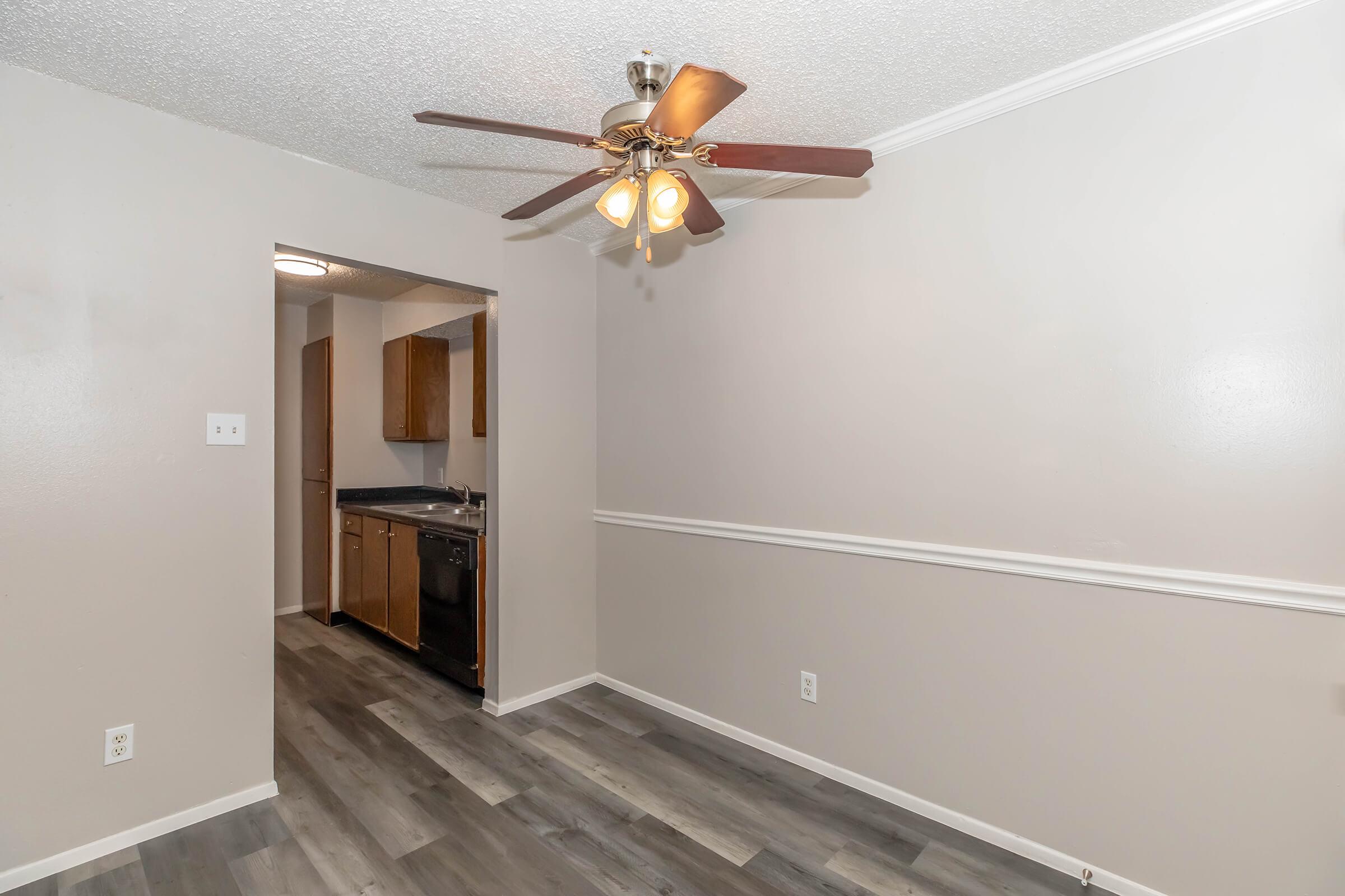 Interior view of a living space featuring a ceiling fan with wooden blades, light-colored walls, and vinyl flooring. The room has a doorway leading to a kitchen area with wooden cabinets and appliances visible. The space is well-lit and clean, suggesting a modern, minimalistic design.