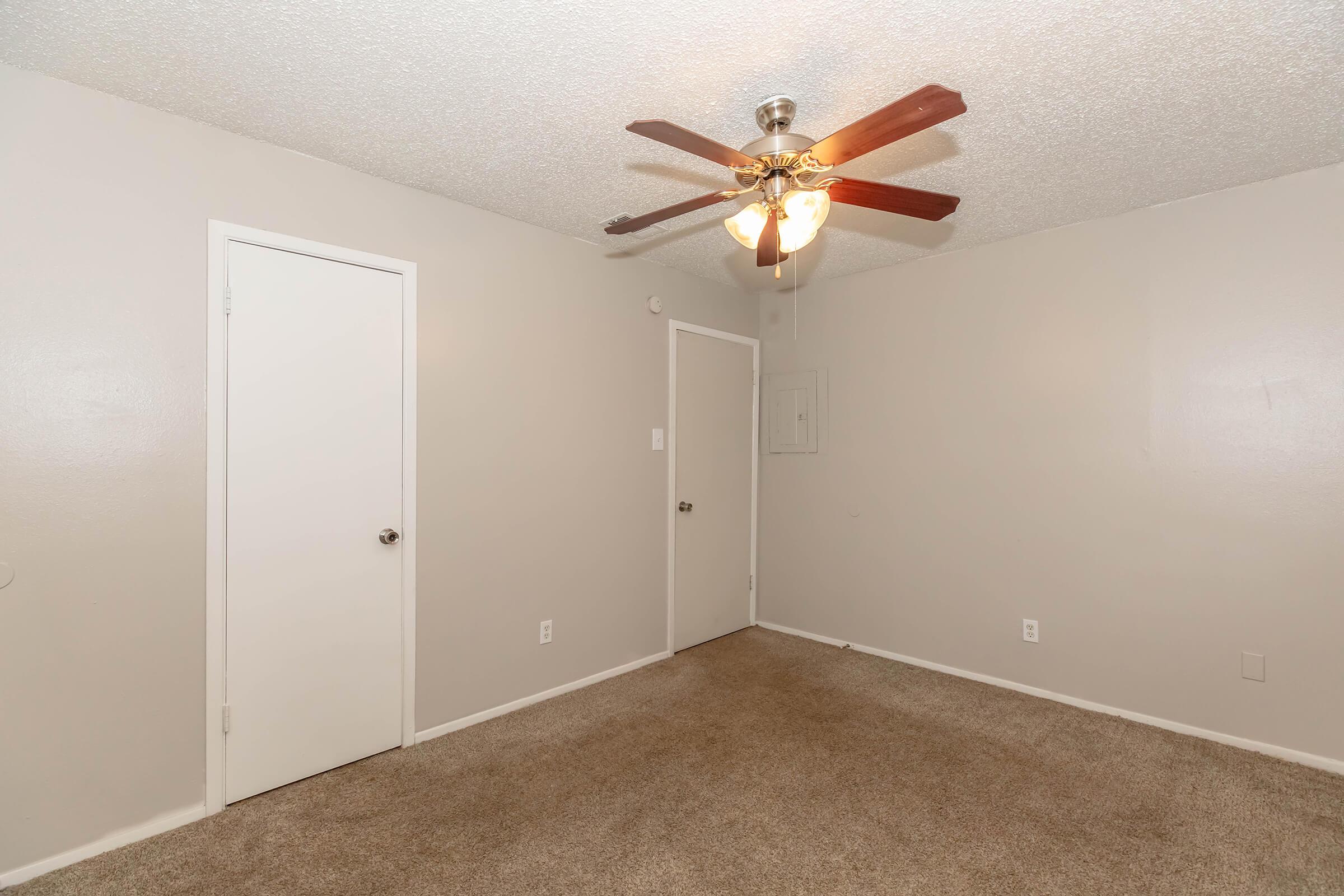 A spacious room featuring light beige carpet, a ceiling fan with wooden blades, and two doors. The walls are painted in a soft grey tone, giving the room a neutral and bright atmosphere. Ideal for various uses, such as a bedroom or office.