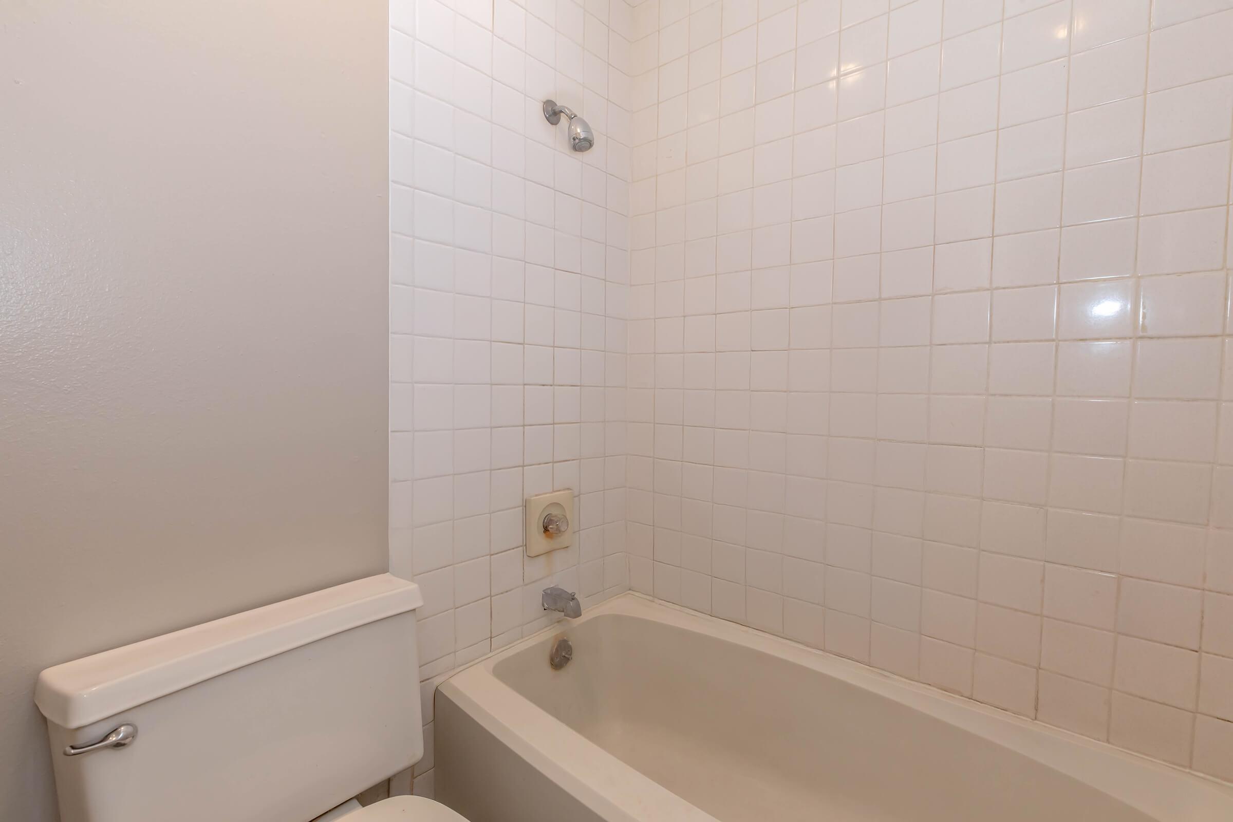A clean, minimalist bathroom featuring a white tiled wall, a bathtub with a faucet, and a toilet. The space is well-lit, highlighting the simple design and neutral colors of the fixtures.