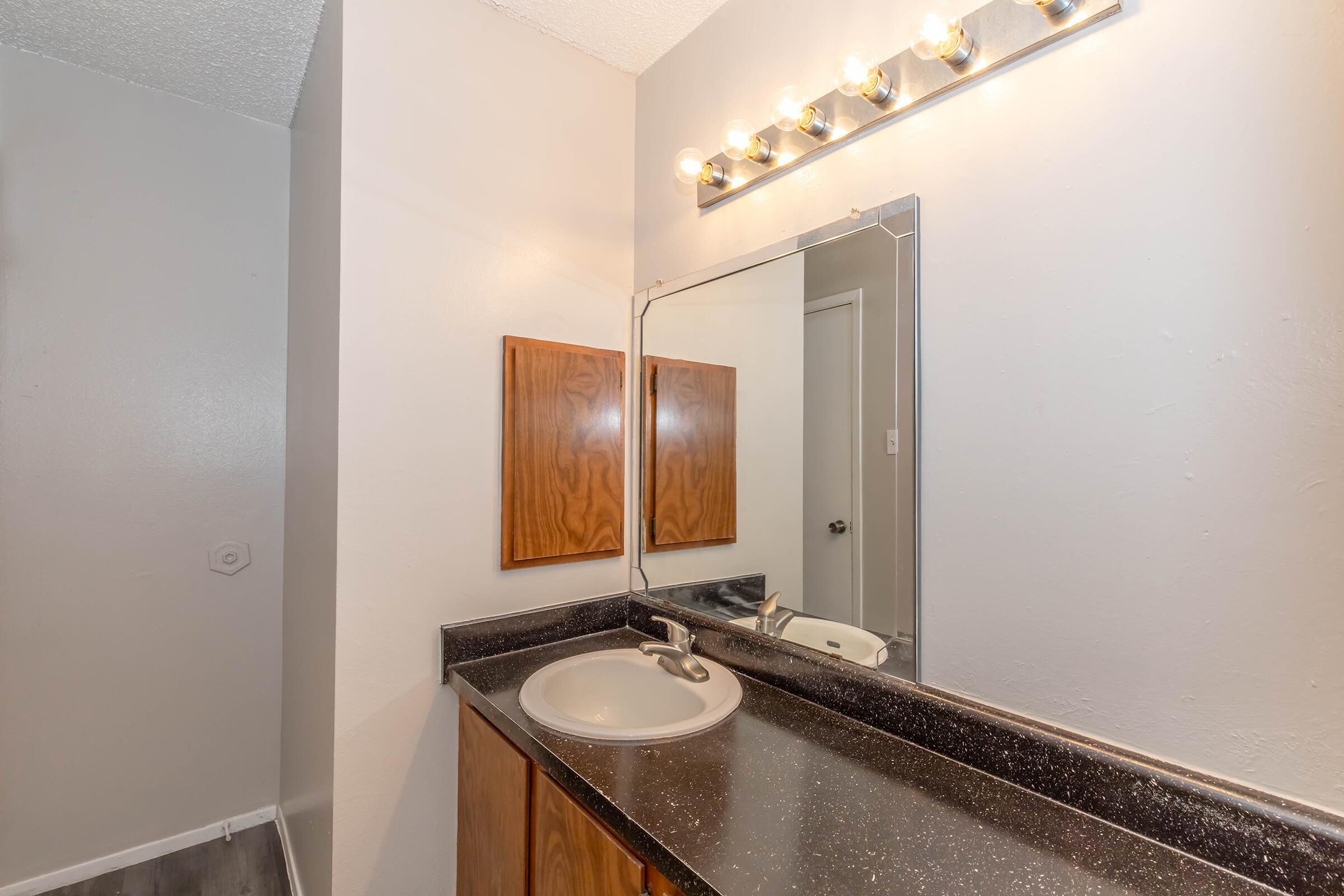 Bathroom setting featuring a simple countertop with a sink, a large mirror above, and wall-mounted light fixtures. Two wooden-framed panels are mounted next to the mirror. The wall is painted in a light shade, and the floor has a light gray finish. The overall design is clean and functional.