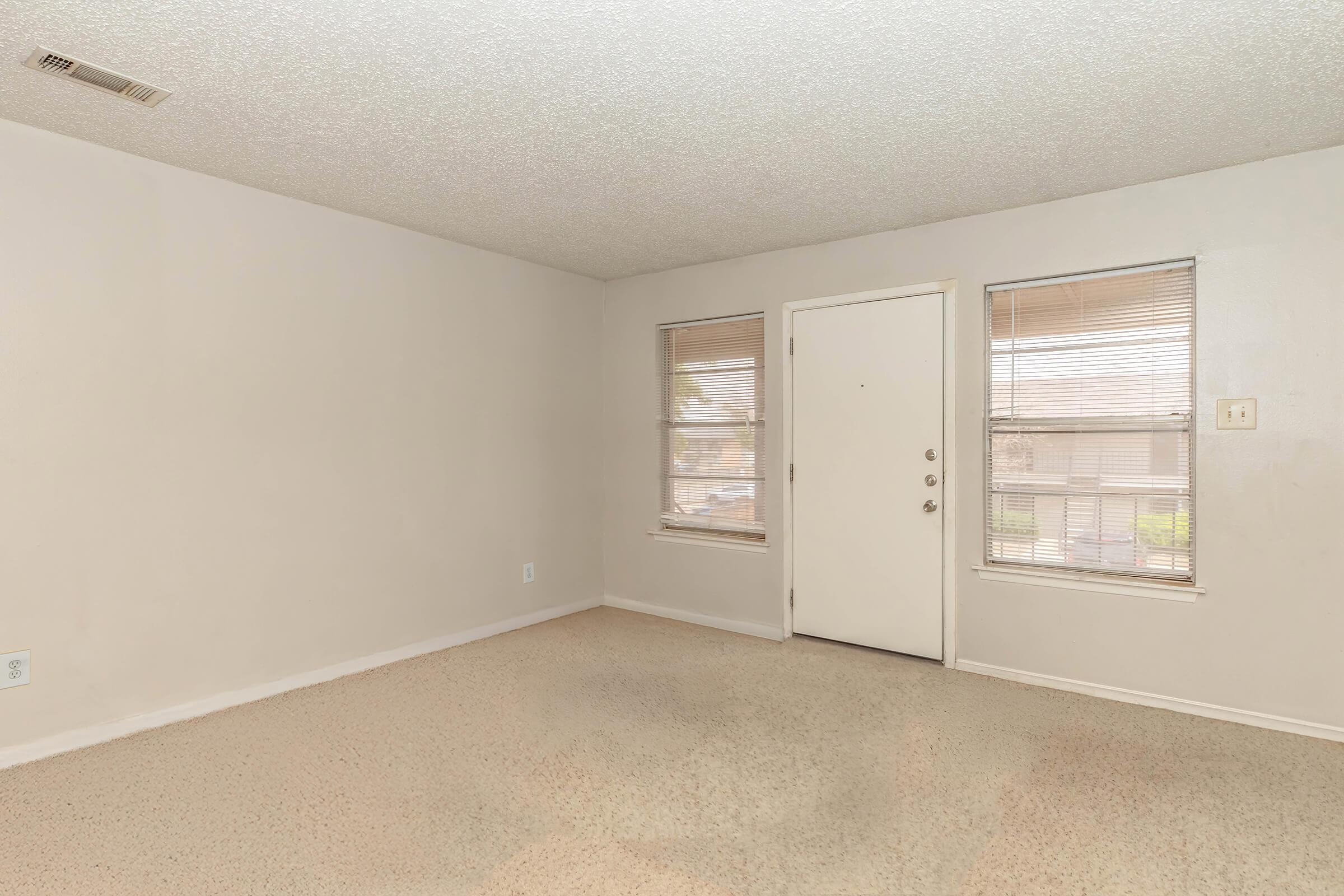 A small, empty room with beige carpet and light gray walls. It features two windows with blinds, a white door, and a plain ceiling. The space is well-lit, creating a neutral and open atmosphere, ideal for various uses or furnishings.
