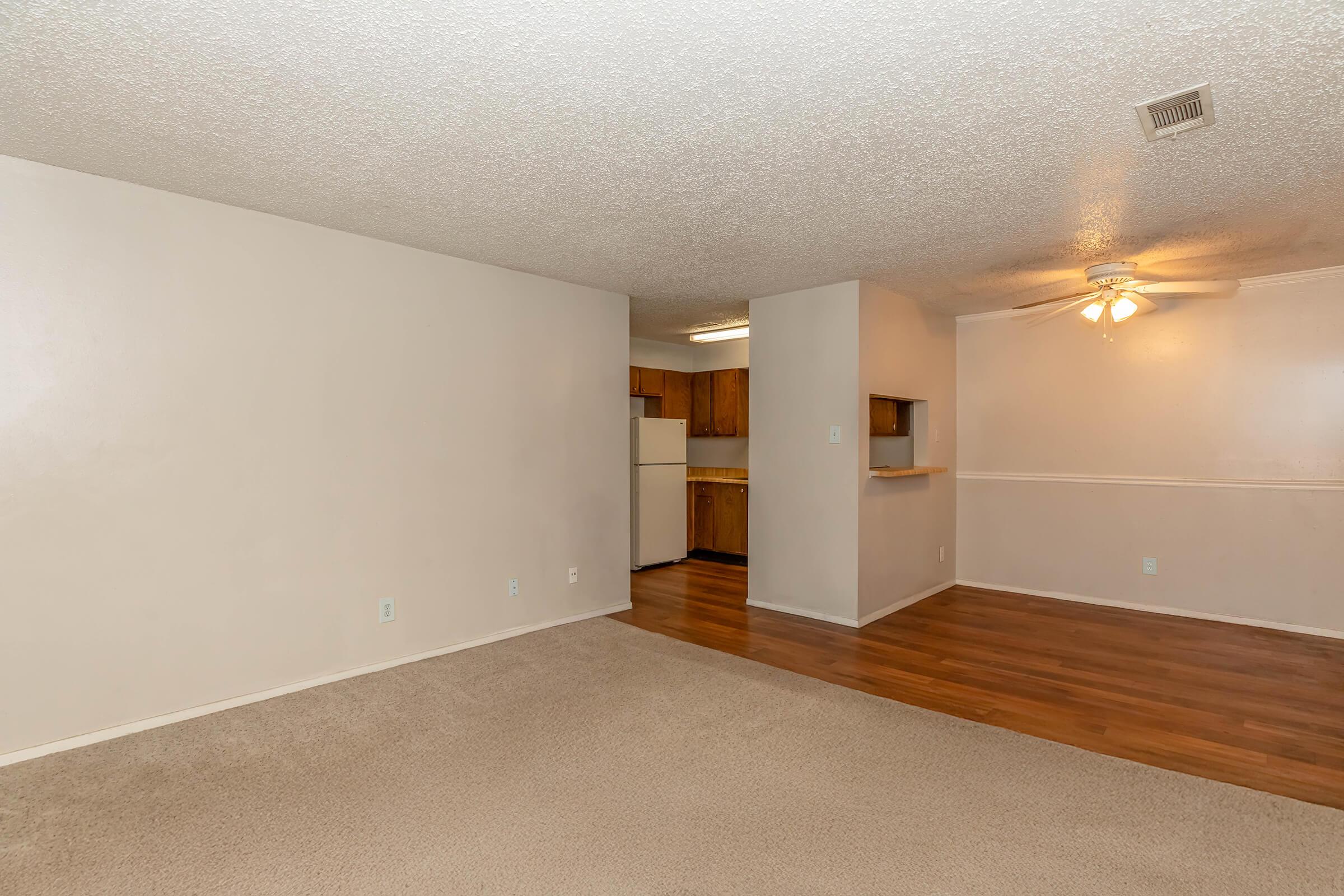 Spacious living area with light-colored walls and carpet. A small kitchen area is visible through an open doorway, featuring wooden cabinetry and a white refrigerator. A ceiling fan is mounted in the corner of the room. The flooring transitions from carpet to wood. Natural light brightens the space.