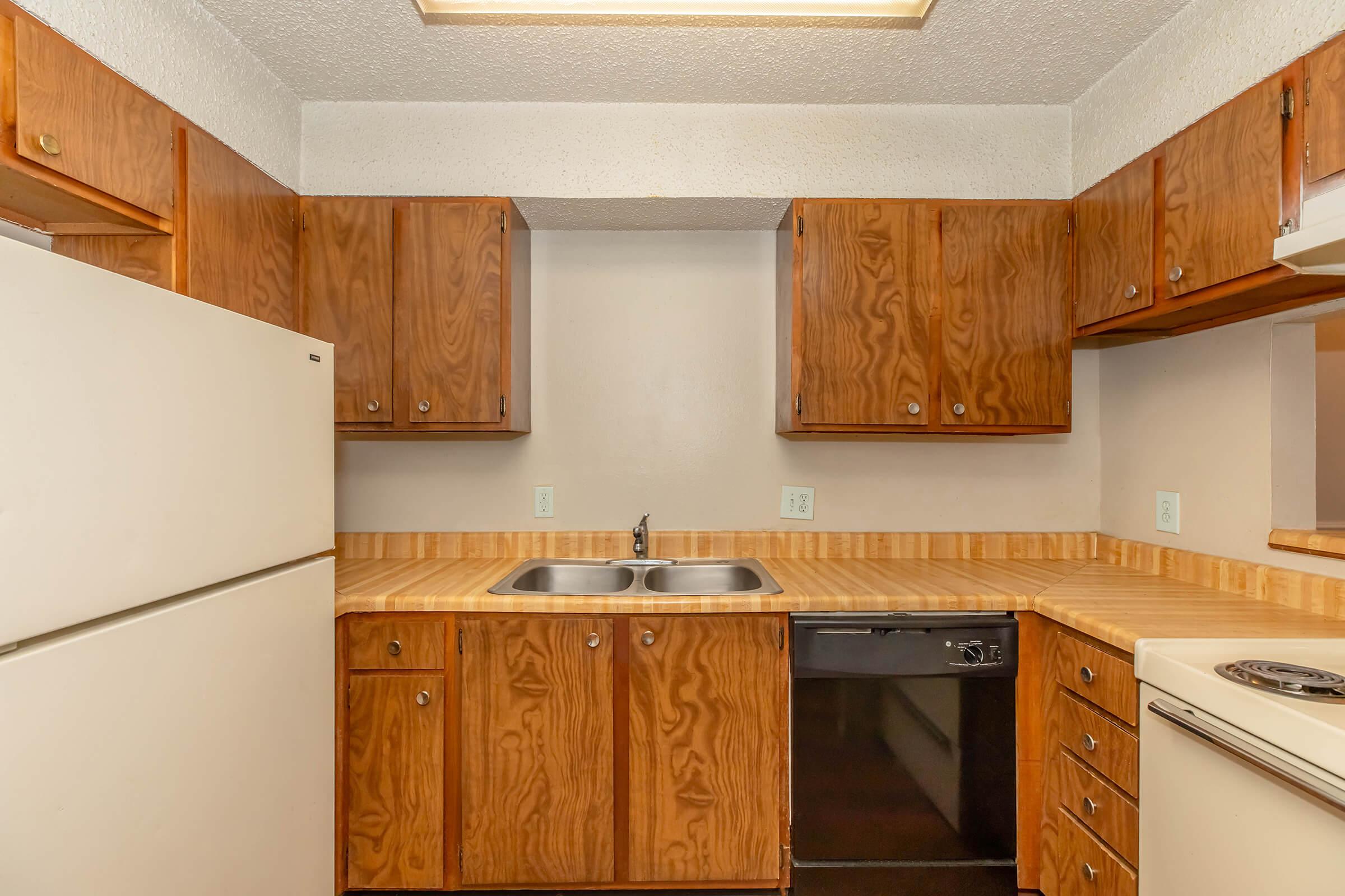A small kitchen featuring wooden cabinets, a double sink, a black dishwasher, a white refrigerator, and a beige countertop. The walls are painted in light colors, and the kitchen has basic appliances arranged in a compact layout. Bright overhead lighting provides illumination.