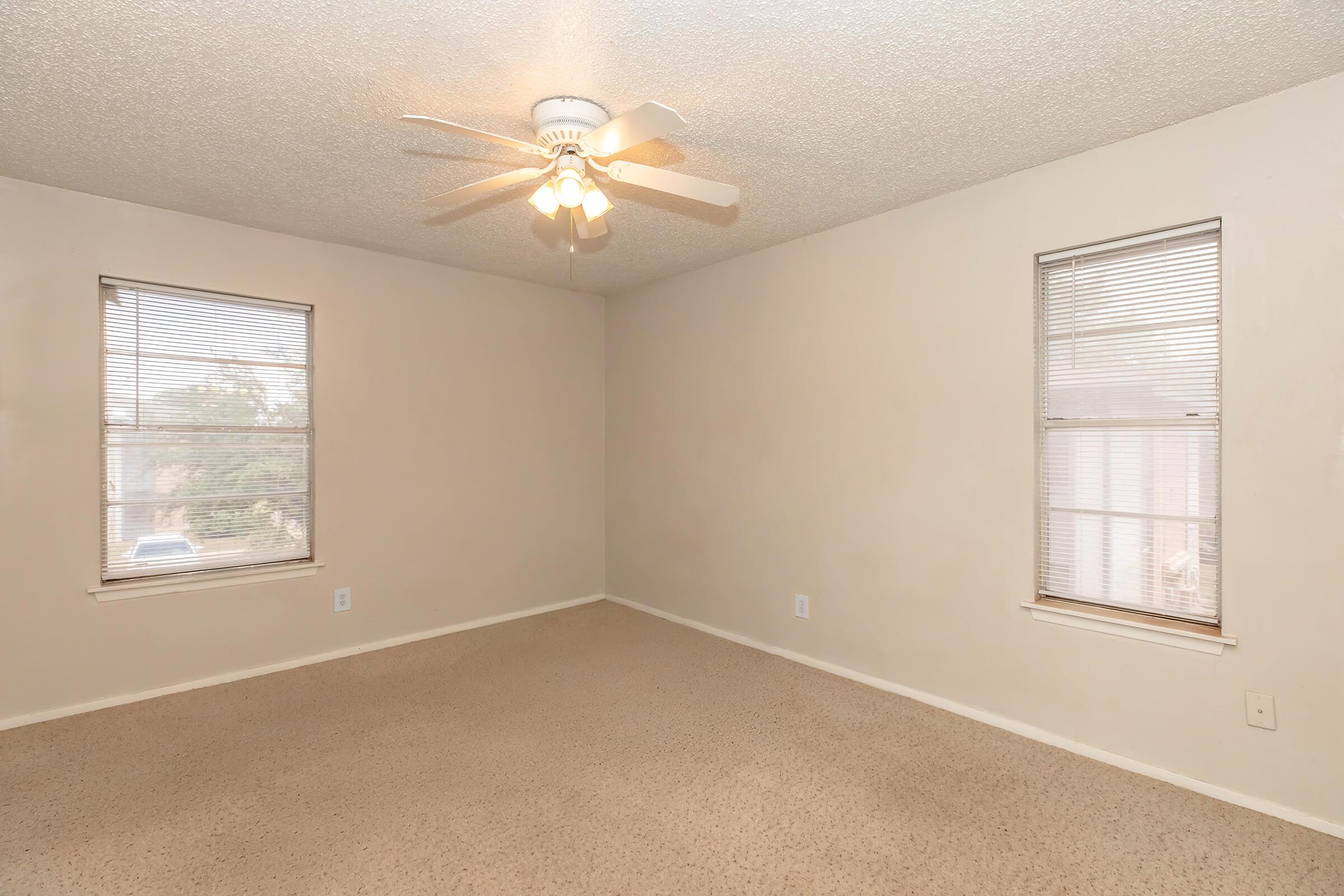 Empty room with light beige walls and a ceiling fan with four blades. The floor is covered in light carpet. Two windows with blinds are installed on opposite walls, allowing natural light to enter the space. The room is uncluttered and ready for decoration.