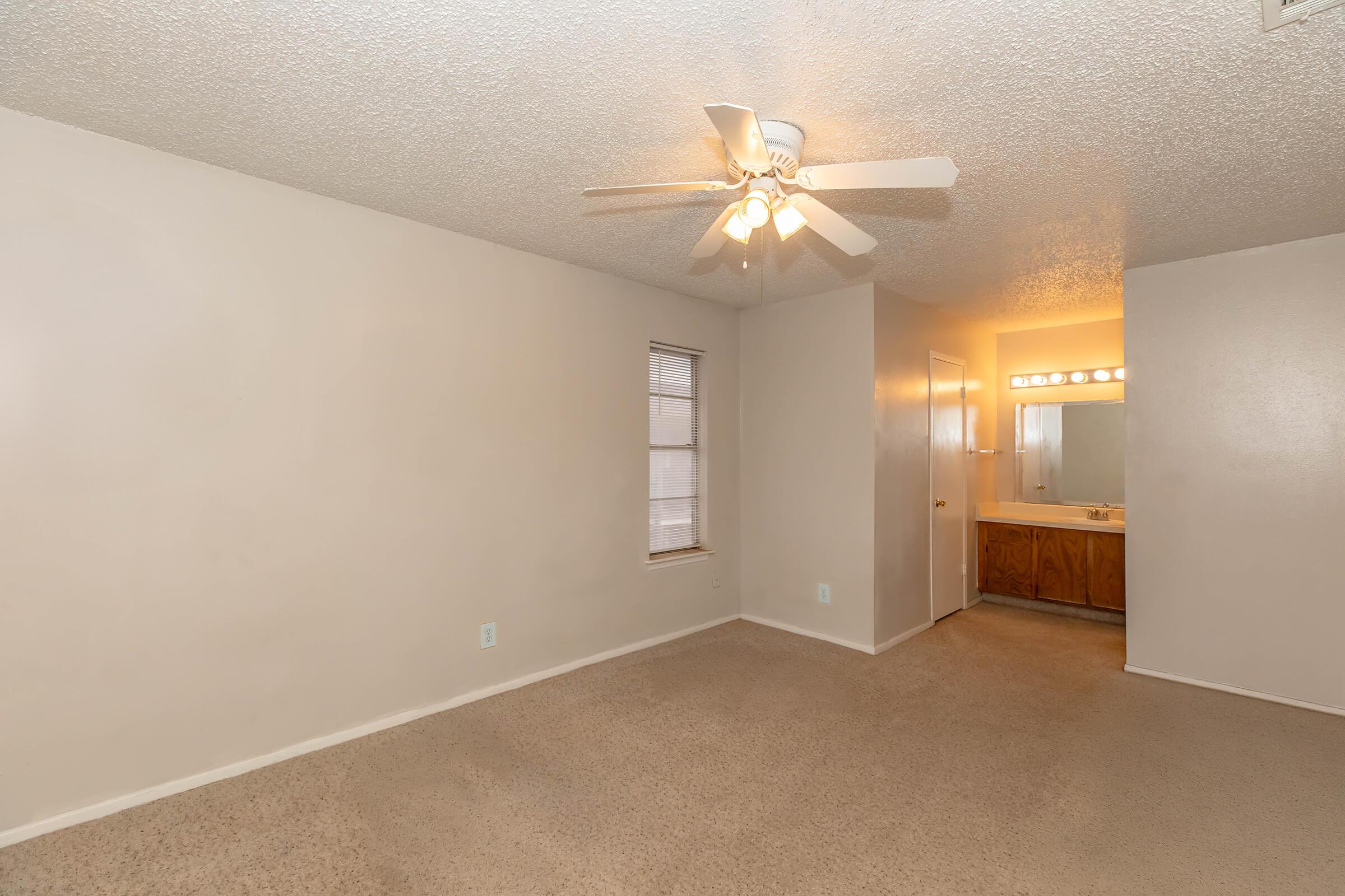 A spacious, neutral-toned room with beige carpet, featuring a ceiling fan and a small window. To the right, a doorway leads to a bathroom with warm lighting and wooden cabinetry. The walls are light-colored, creating an open and inviting atmosphere.