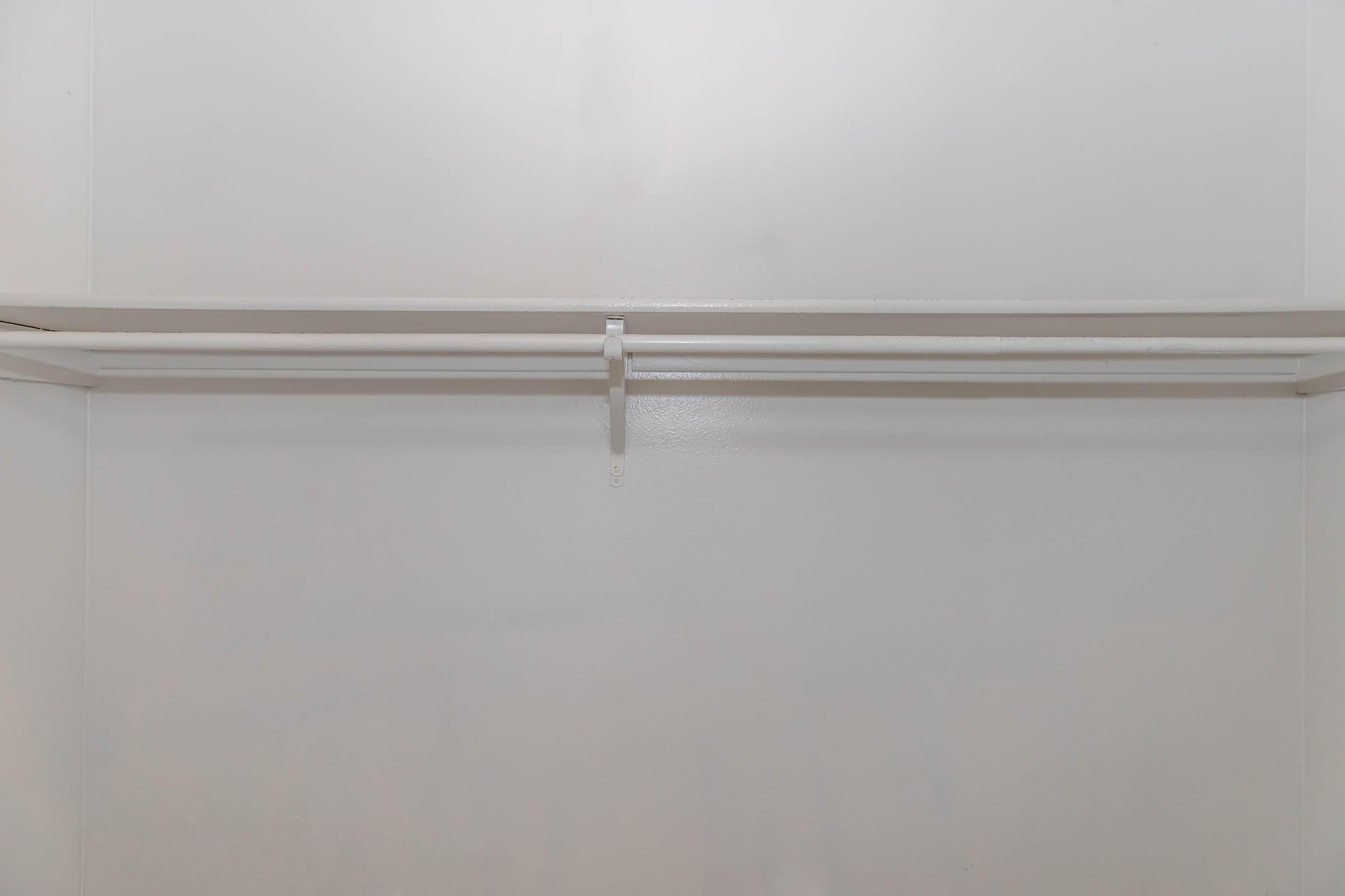 Empty closet interior featuring plain white walls and a single white clothing rod suspended in the middle. The design is minimalistic, with no additional fixtures or items visible, creating a clean and open space.