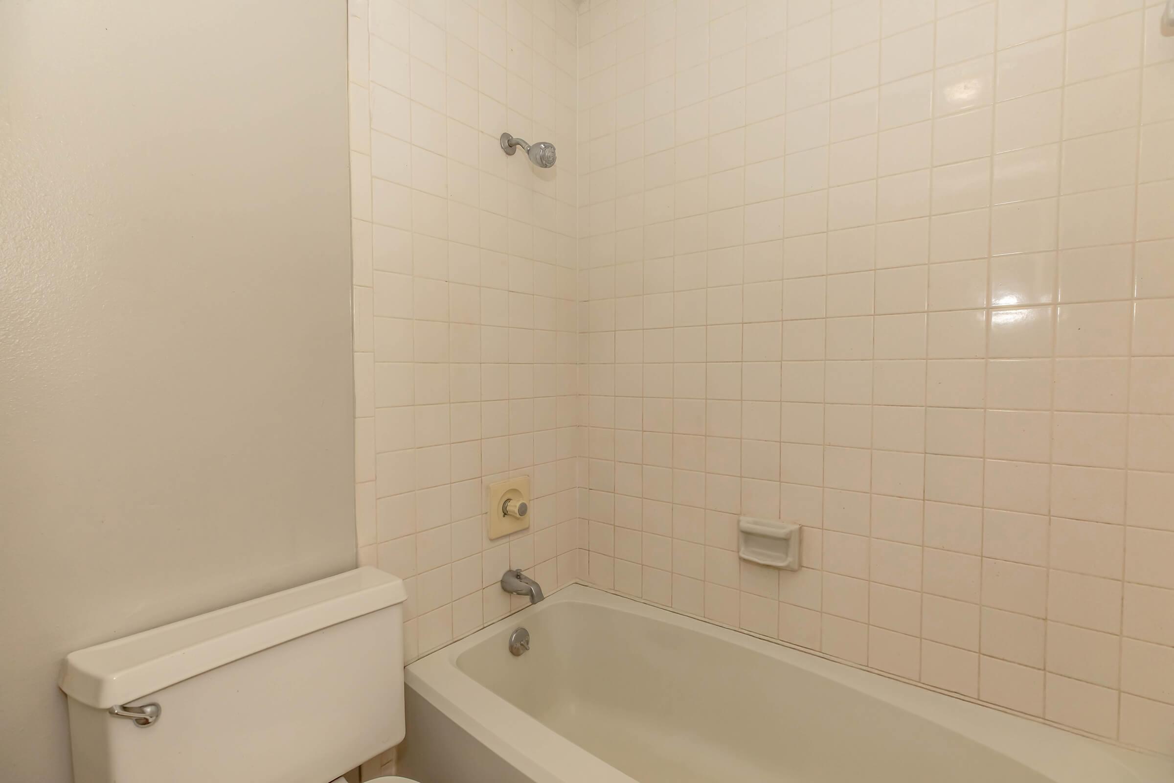 A simple bathroom featuring a white tiled shower area with a bathtub, a showerhead, and a soap dish. To the left, there is a toilet with a low tank. The walls are light-colored, creating a clean and minimalistic look.