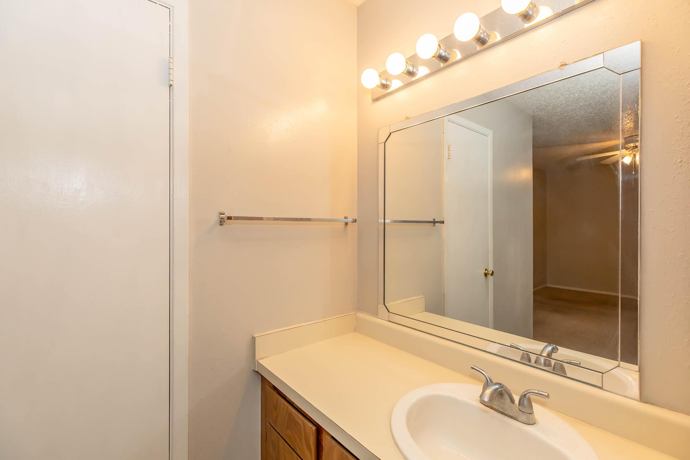 A bathroom with a simple sink and countertop, large mirror with overhead lights, and a towel rack. A door is partially visible on the left, and a neutral-colored wall leads to a room shown in the background. The overall atmosphere is clean and minimalistic.
