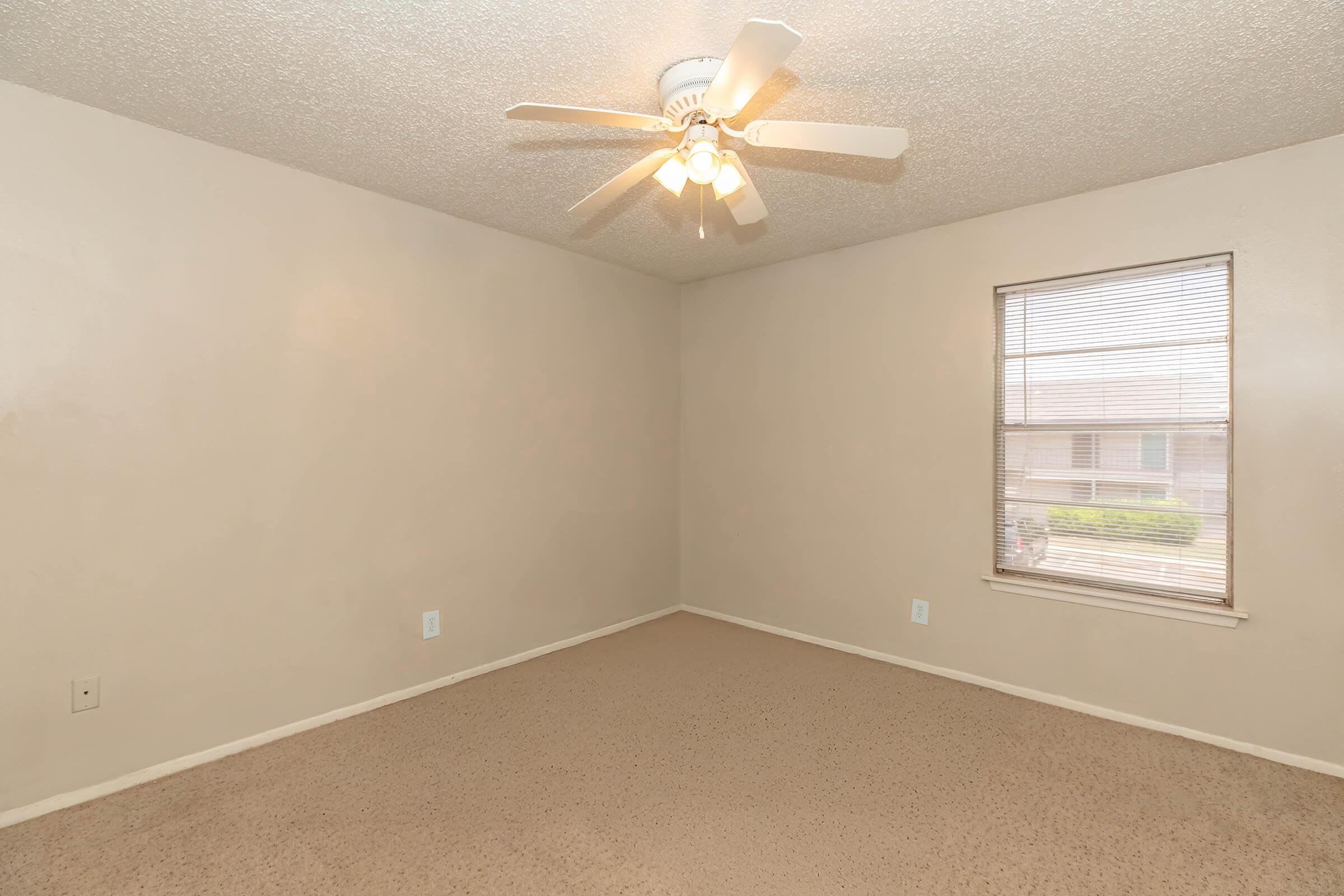 A spacious, empty bedroom with beige walls and carpet. A ceiling fan with four lights hangs from the ceiling. There is a window on the right side allowing natural light to enter, and the room appears clean and ready for furnishing.