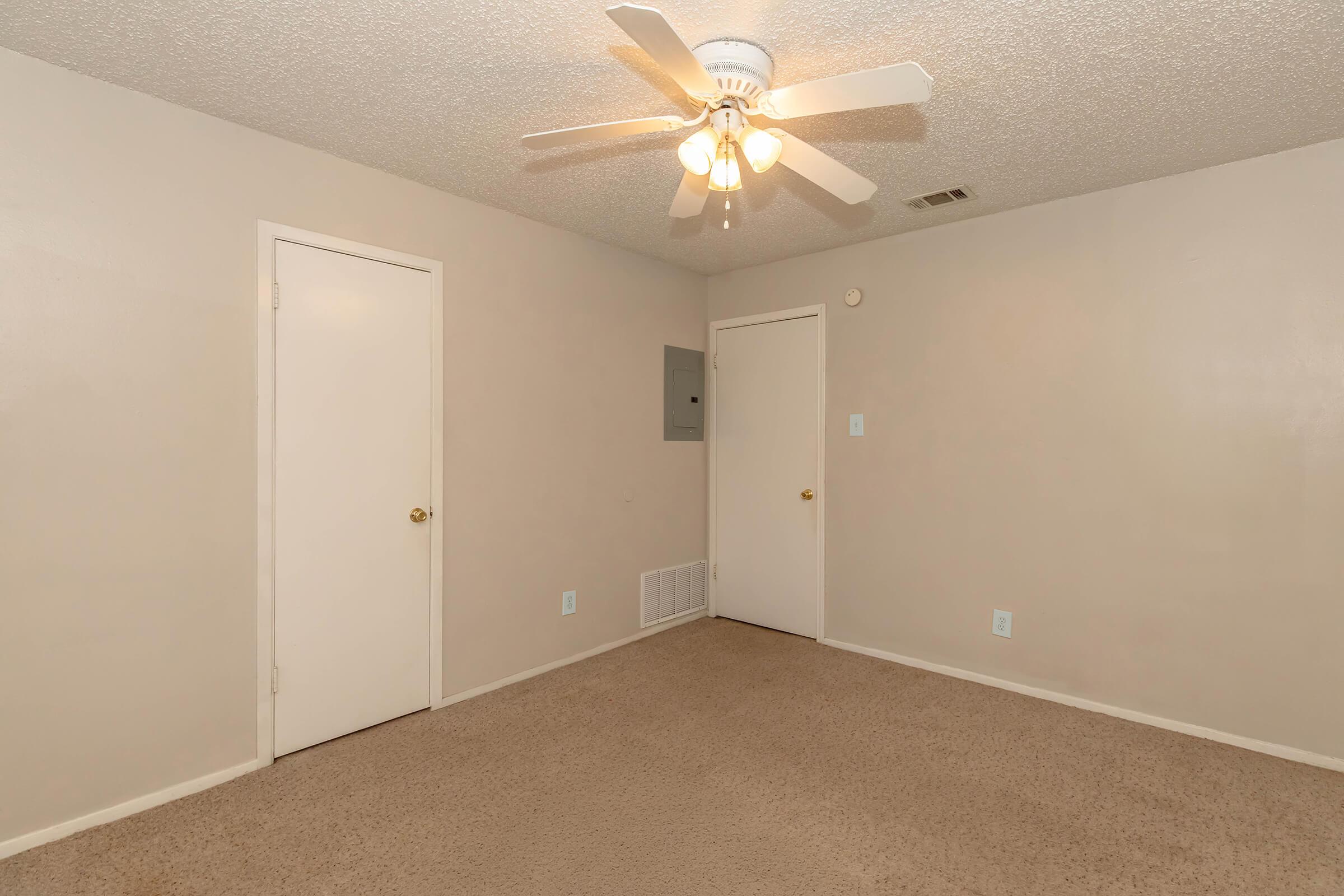 A spacious, empty room with light beige walls and a light brown carpet. There is a ceiling fan with light fixtures in the center. Two doors are visible: one leading to another space and the other possibly a closet or utility area. The room has a clean and simple aesthetic.