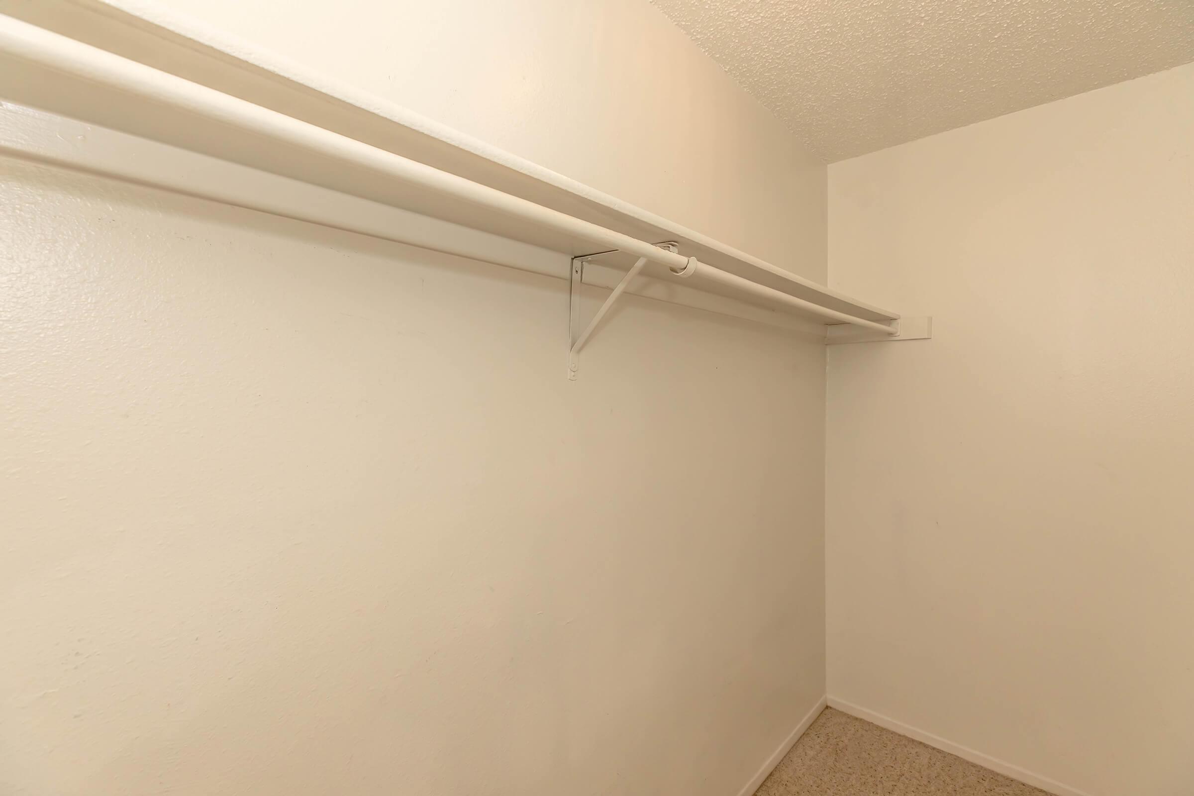 An empty closet with a smooth white wall and a single clothes rod mounted along the top. The floor is covered in light-colored carpeting, and there are no shelves or additional furniture present. The space appears clean and uncluttered, emphasizing its potential for storage.