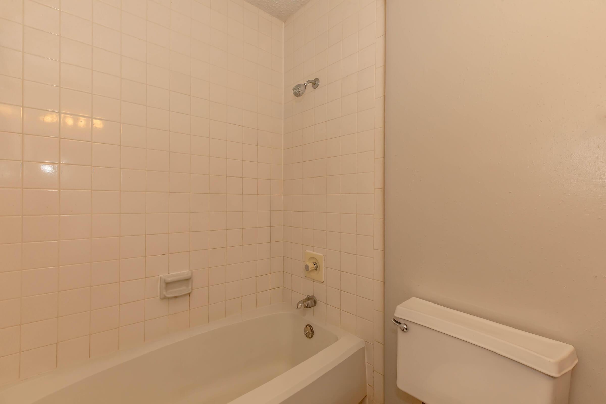 Interior view of a bathroom featuring a white bathtub against a beige tiled wall. The showerhead is mounted above the tub, and a light switch is visible on the wall. A toilet is positioned to the right, also in white. The space is well-lit and minimalistic, with neutral color tones.