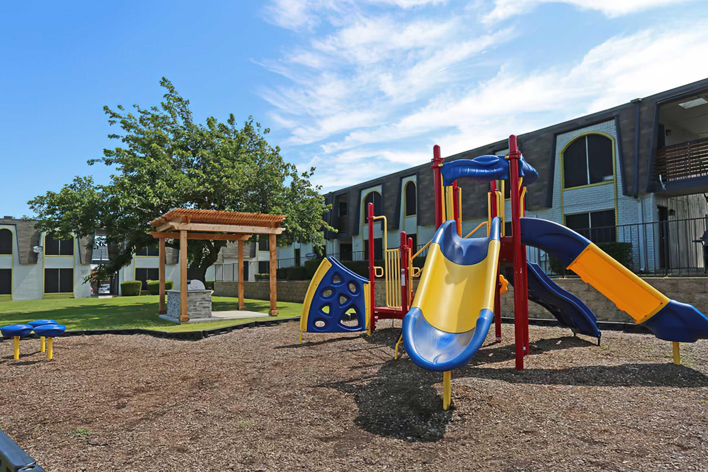 A colorful children's playground featuring yellow and blue slides, climbing structures, and a wooden gazebo. The area is surrounded by green grass and trees, with an apartment building in the background under a clear blue sky.