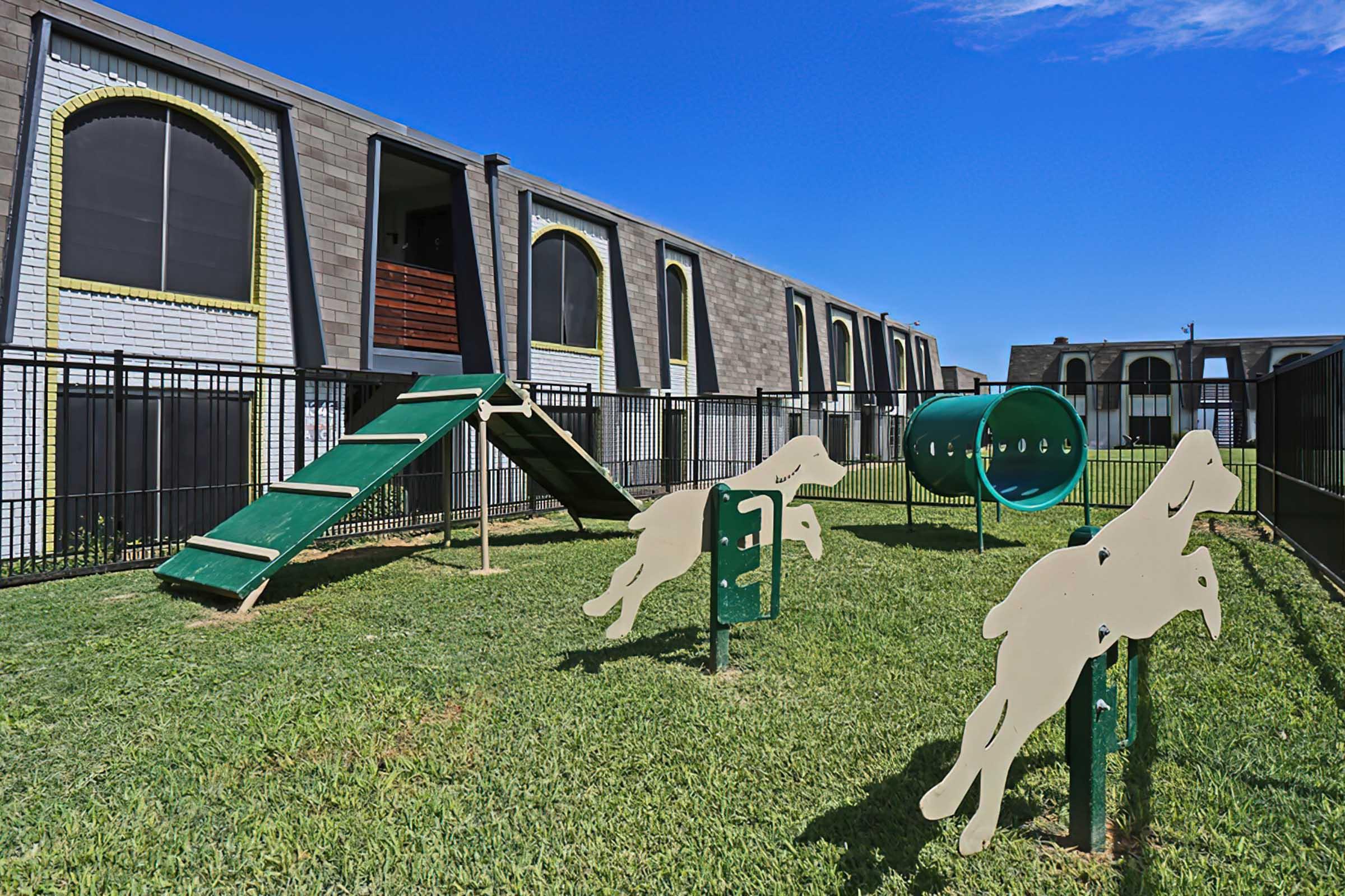 A fenced dog play area featuring toys and equipment, including a small incline ramp and a tunnel, surrounded by well-maintained grass and adjacent to modern buildings. The sky is clear with a few clouds visible.