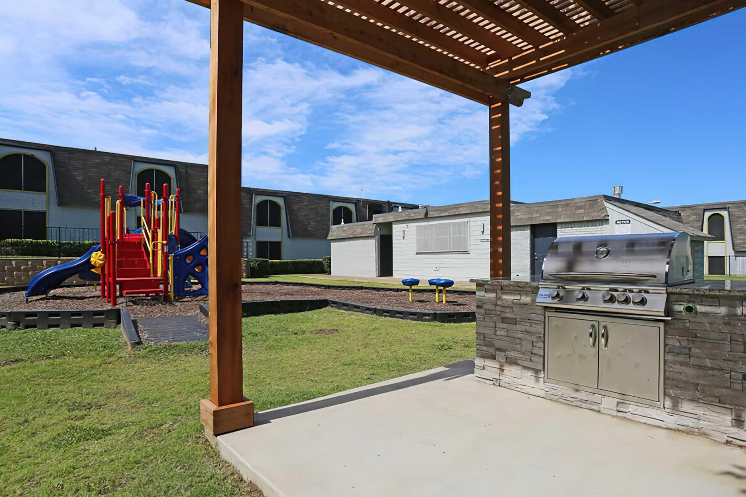 A family-friendly outdoor space featuring a playground with colorful equipment, picnic area with a gas grill under a wooden pergola, and a grassy area. The background includes a building with multiple units, creating a welcoming atmosphere for gatherings and play.