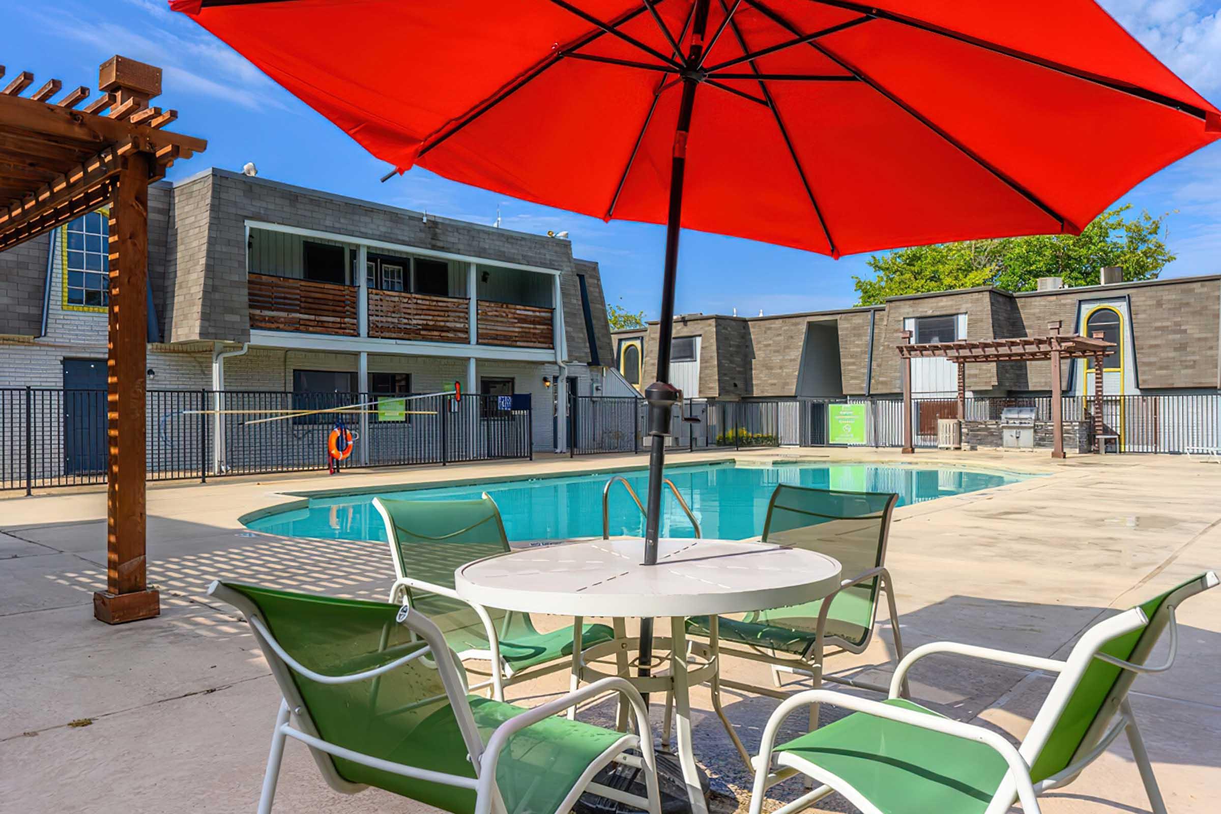 A swimming pool area featuring a vibrant red umbrella over a round table with green chairs. Surrounding the pool are apartment buildings with a mix of modern and traditional designs. A safety buoy is visible near the water, and the sky is clear and sunny, creating a relaxing outdoor atmosphere.