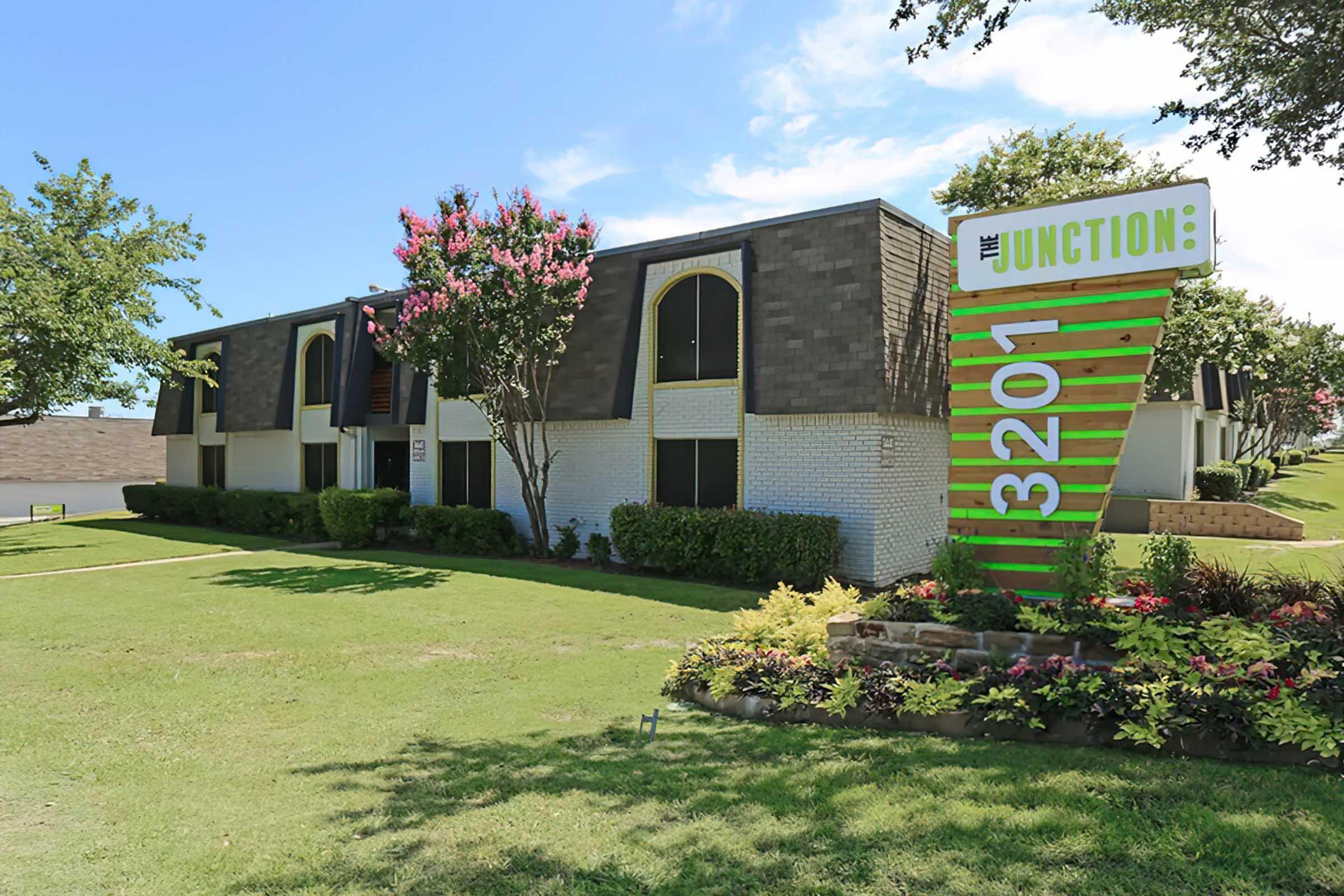 A modern apartment complex with a green and white sign displaying "Junction" and the address "3201." The building features dark roofs and large windows, surrounded by well-maintained lawns and landscaping with colorful flowers and shrubs under a clear blue sky.