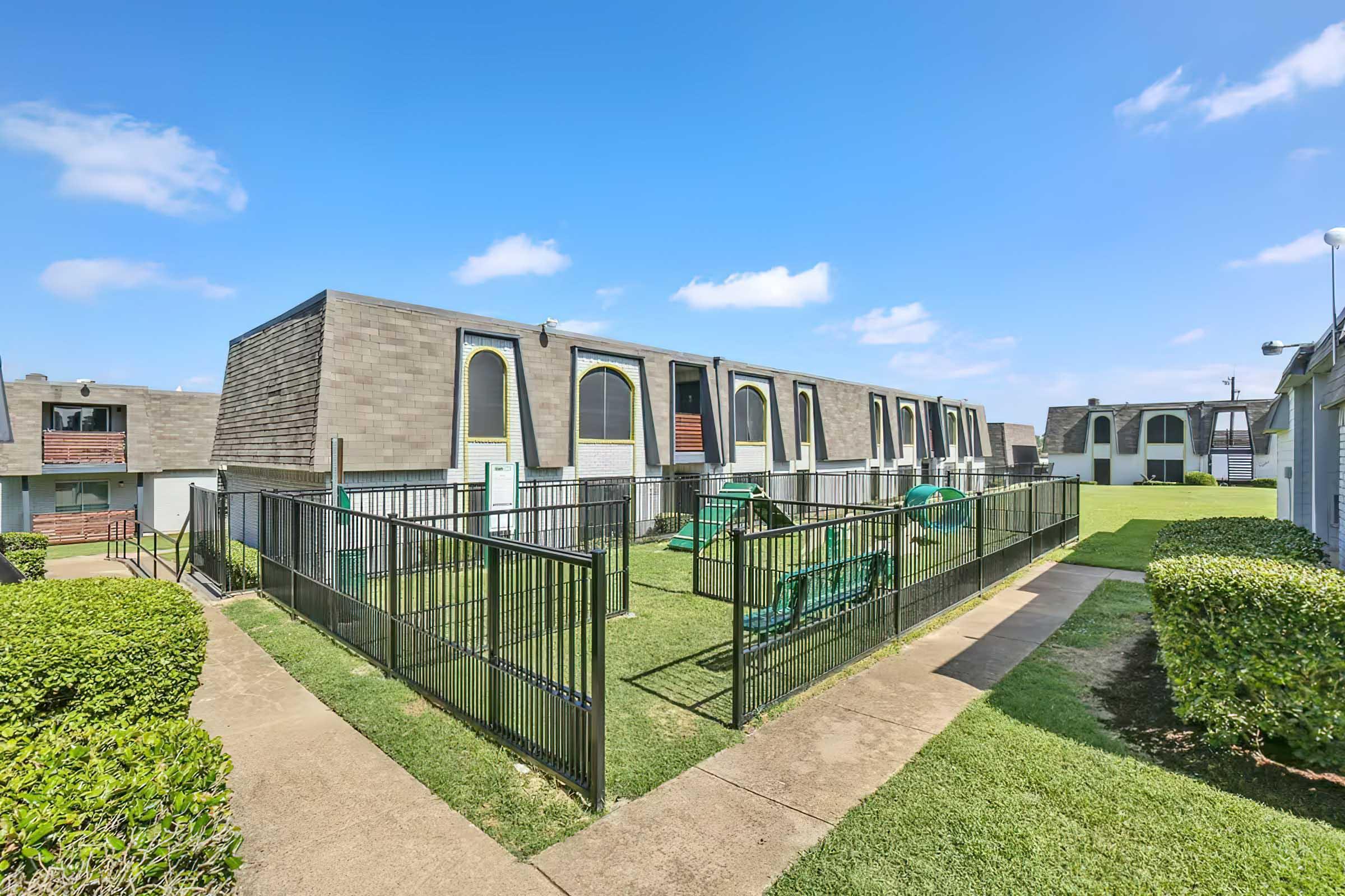 A view of a modern apartment complex featuring several units with distinct window shapes. The grounds have well-maintained green grass and a fenced area with playground equipment, surrounded by neatly trimmed hedges. Bright blue sky with a few clouds adds to the pleasant atmosphere.