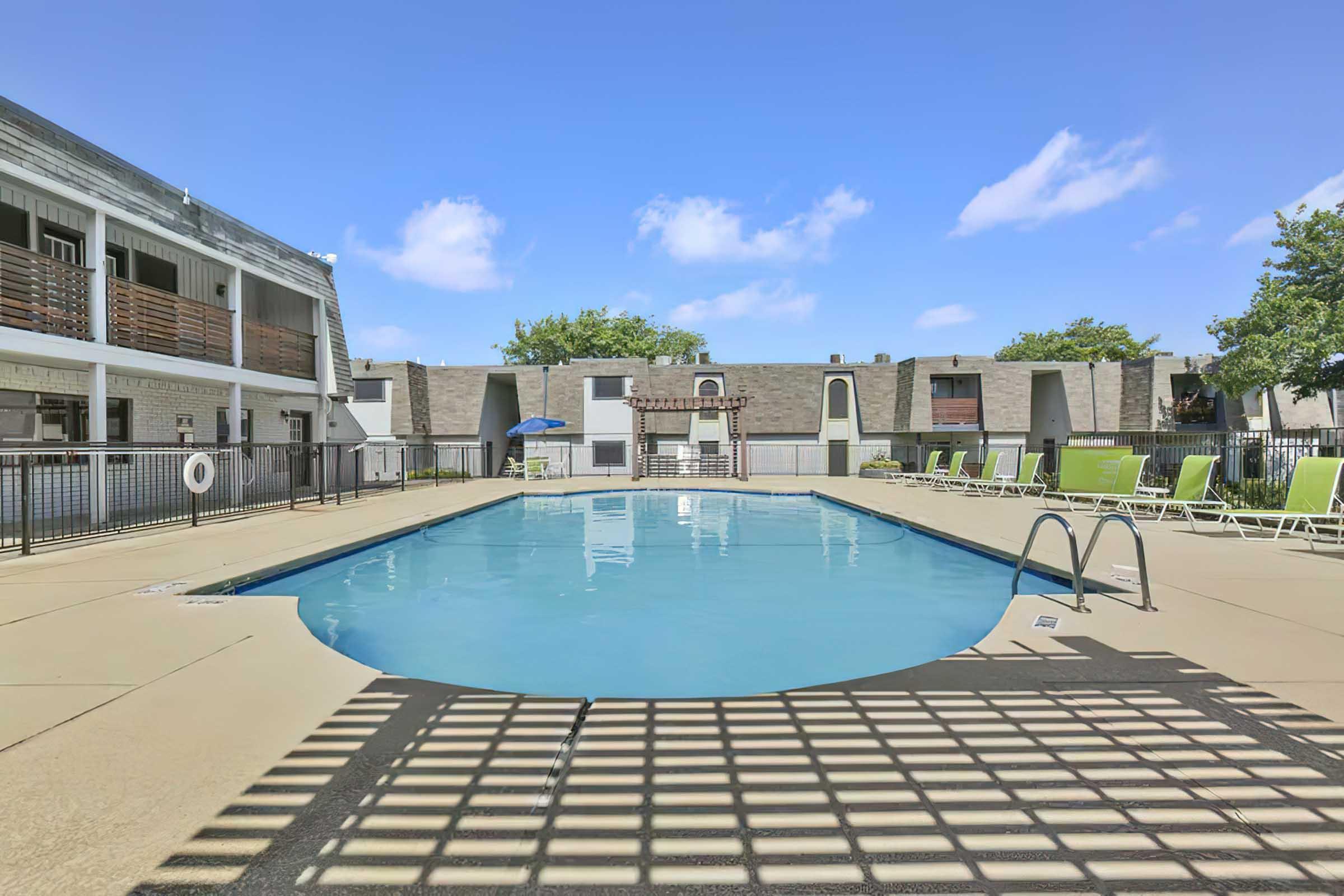A clean outdoor swimming pool surrounded by lounge chairs and shaded areas. The pool features a gradual entry and is enclosed by a fence. In the background, apartment buildings with balconies are visible under a clear blue sky.