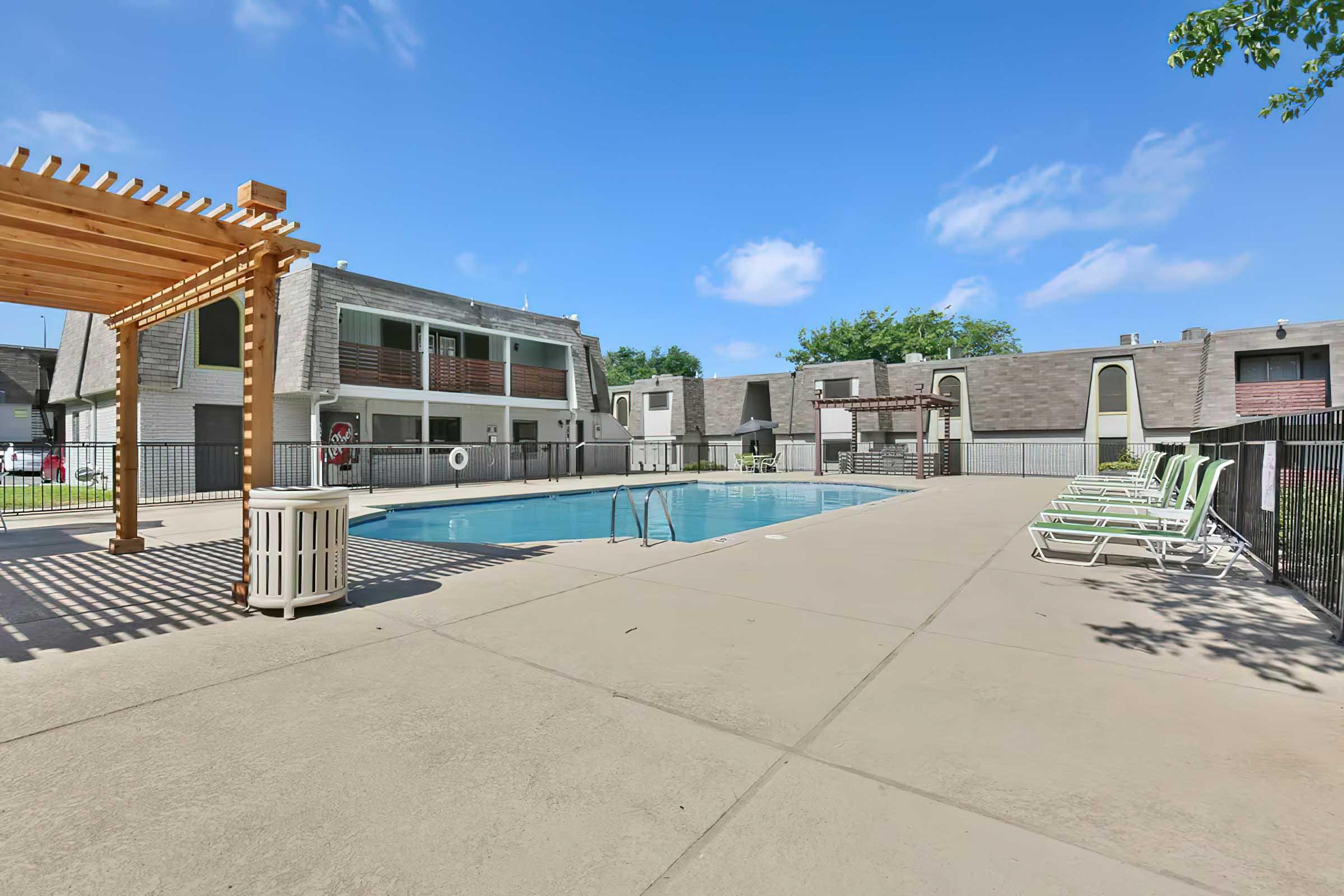 A clear view of a swimming pool surrounded by lounge chairs and a pergola. The pool is set in a residential area with two-story buildings in the background. The sky is bright and blue, creating a welcoming atmosphere for relaxation and enjoyment.