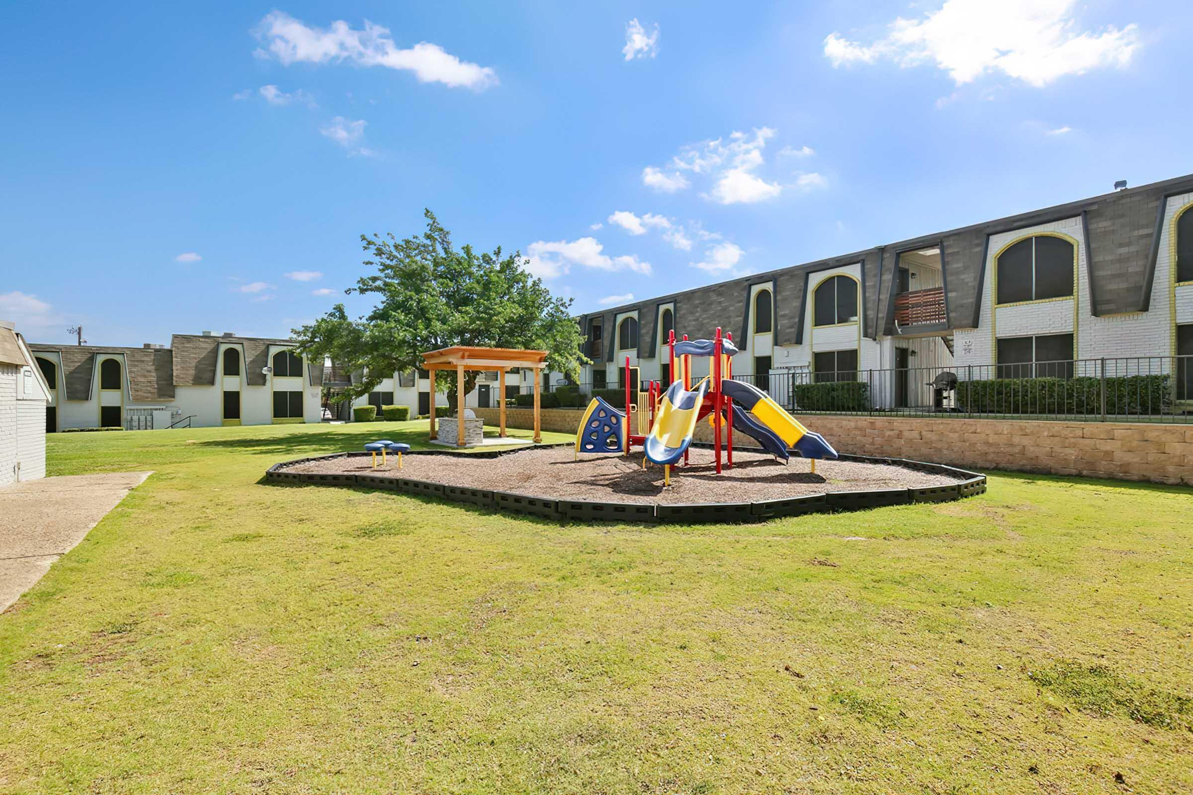 A sunny playground featuring a colorful play structure with slides and climbing equipment, surrounded by green grass. In the background, there are apartment buildings with large windows and a tree providing shade. The sky is bright with a few clouds.