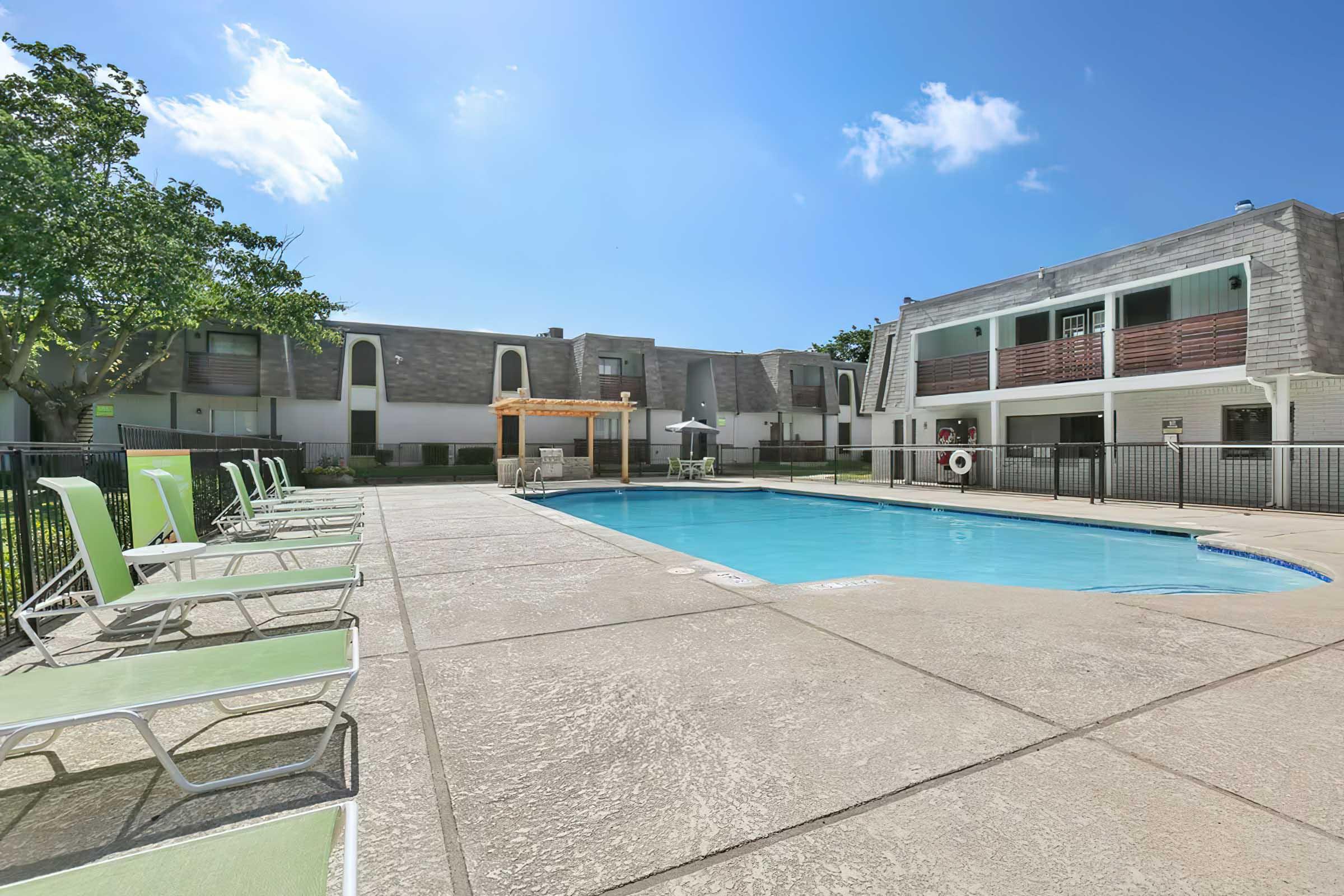 A clear blue swimming pool surrounded by a sunbathing area with green lounge chairs. On one side, there are trees providing shade, and in the background, low-rise apartment buildings with balconies are visible under a bright blue sky.