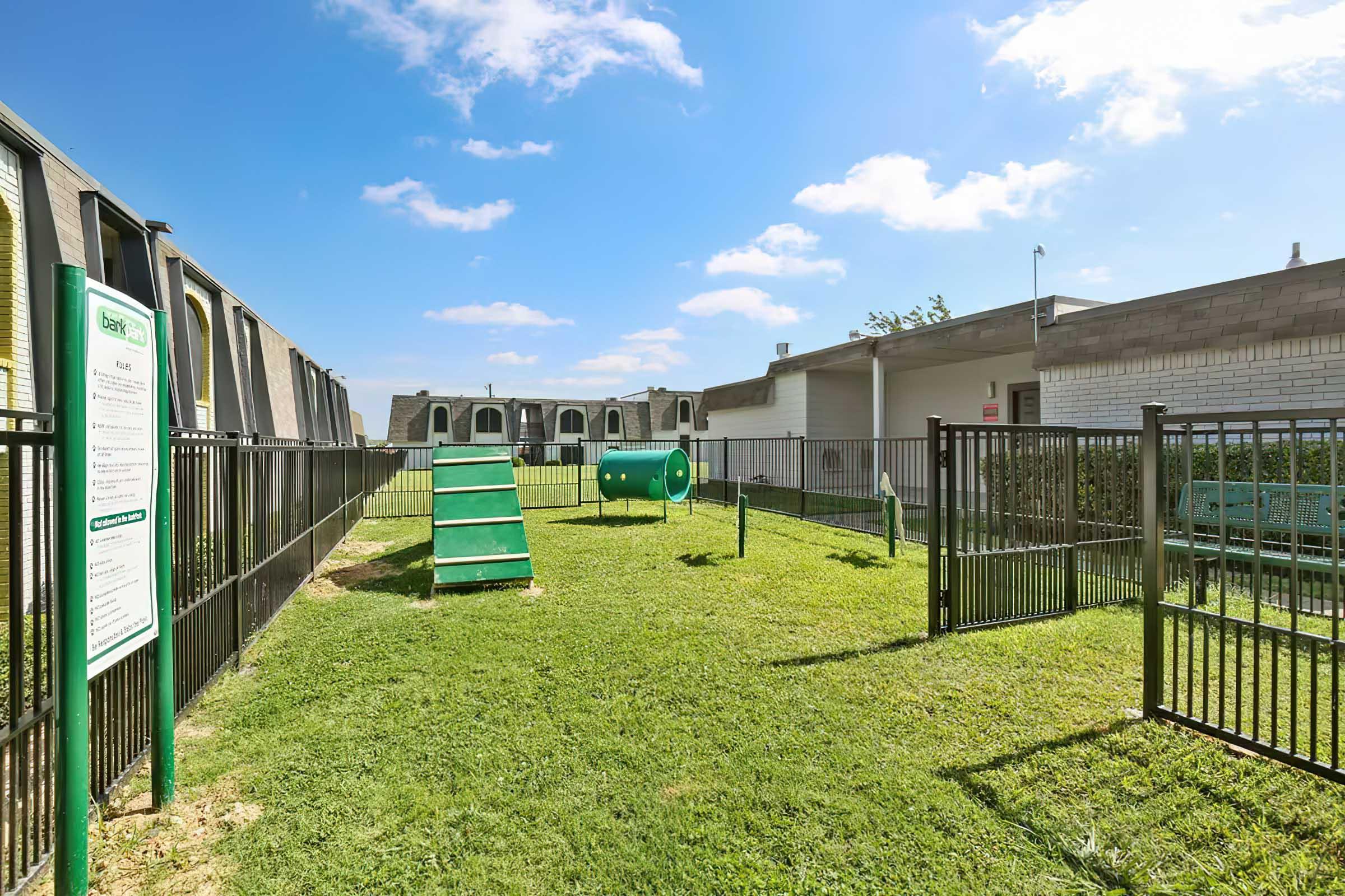 A fenced outdoor area featuring green grass, a slide, and a play tunnel for pets. In the background, there are several buildings and a signpost with information about the facility. The scene is under a blue sky with a few clouds.