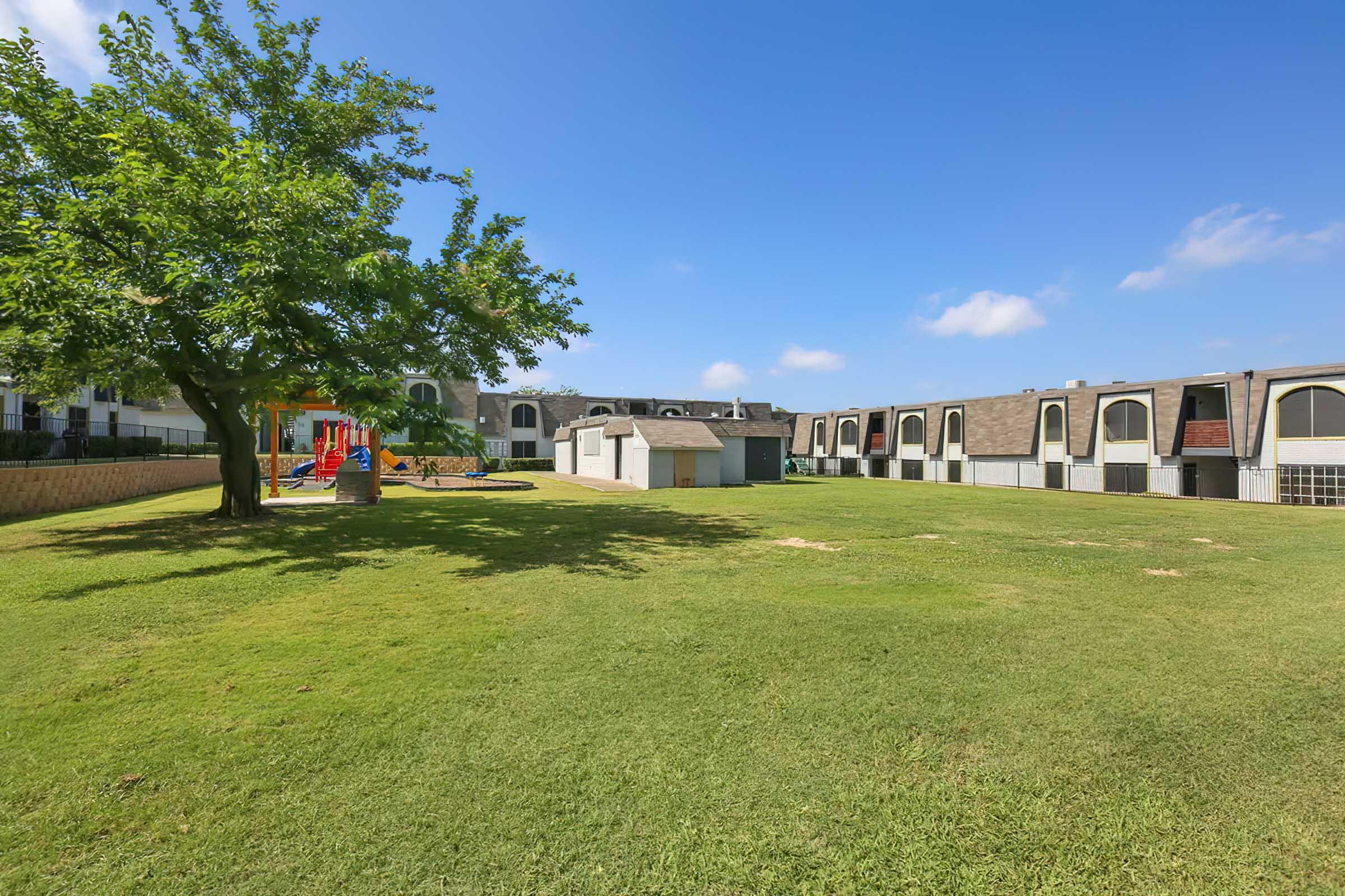 A grassy area featuring a large tree, play structure, and a small shed. In the background, there are buildings with multiple windows under a blue sky with a few clouds. The scene is inviting and well-maintained, perfect for outdoor activities.