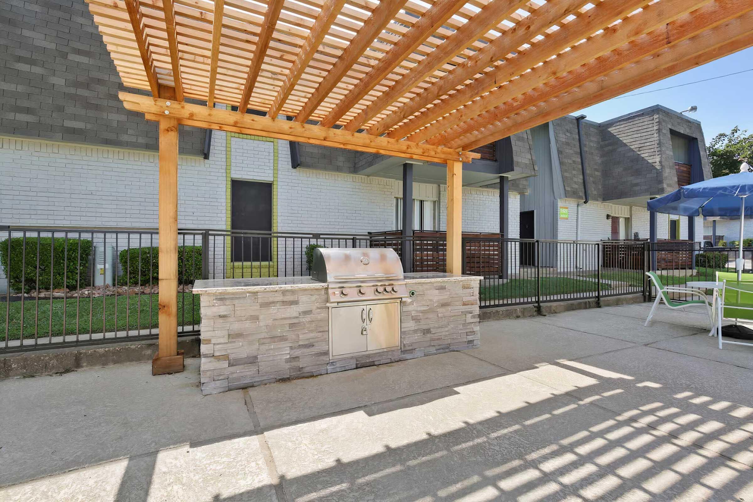 Outdoor grilling area featuring a stone BBQ grill under a wooden pergola, with shaded seating options and a fence in the background. Nearby green landscaping and an apartment complex visible in the distance on a sunny day.