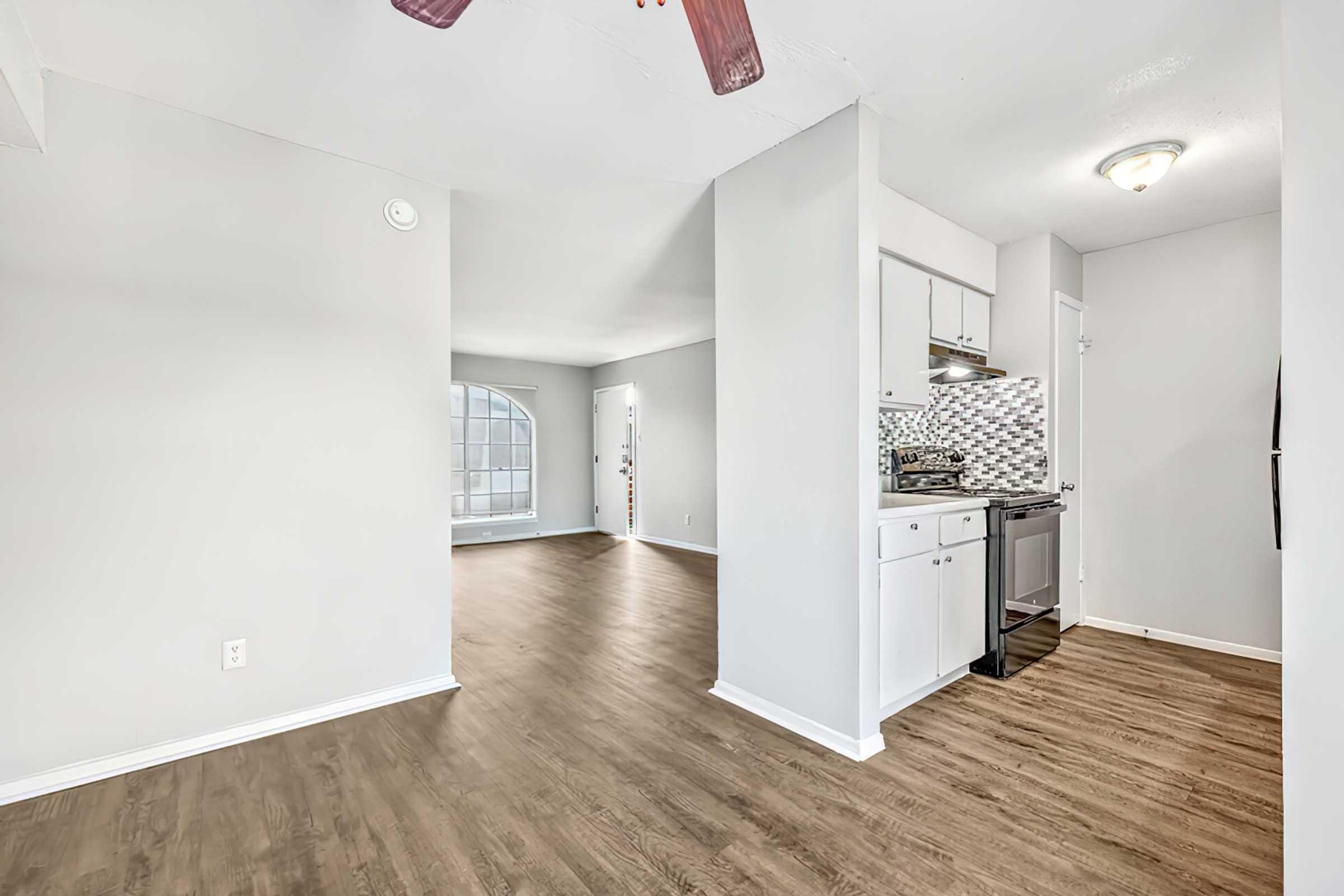 A bright, open living space featuring light gray walls and a modern kitchen. The kitchen includes stainless steel appliances and a tiled backsplash. The flooring is a warm wood finish. Natural light enters through a large window, and a doorway leads to another room.