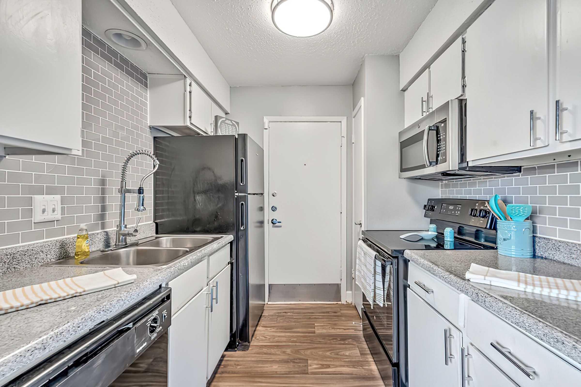 Modern kitchen featuring stainless steel appliances, including a microwave and stove. The countertops are gray with a matching backsplash, and there is a double sink with a stylish faucet. White cabinetry provides storage, and the floor is wooden. A door leads outside, creating a bright, welcoming atmosphere.