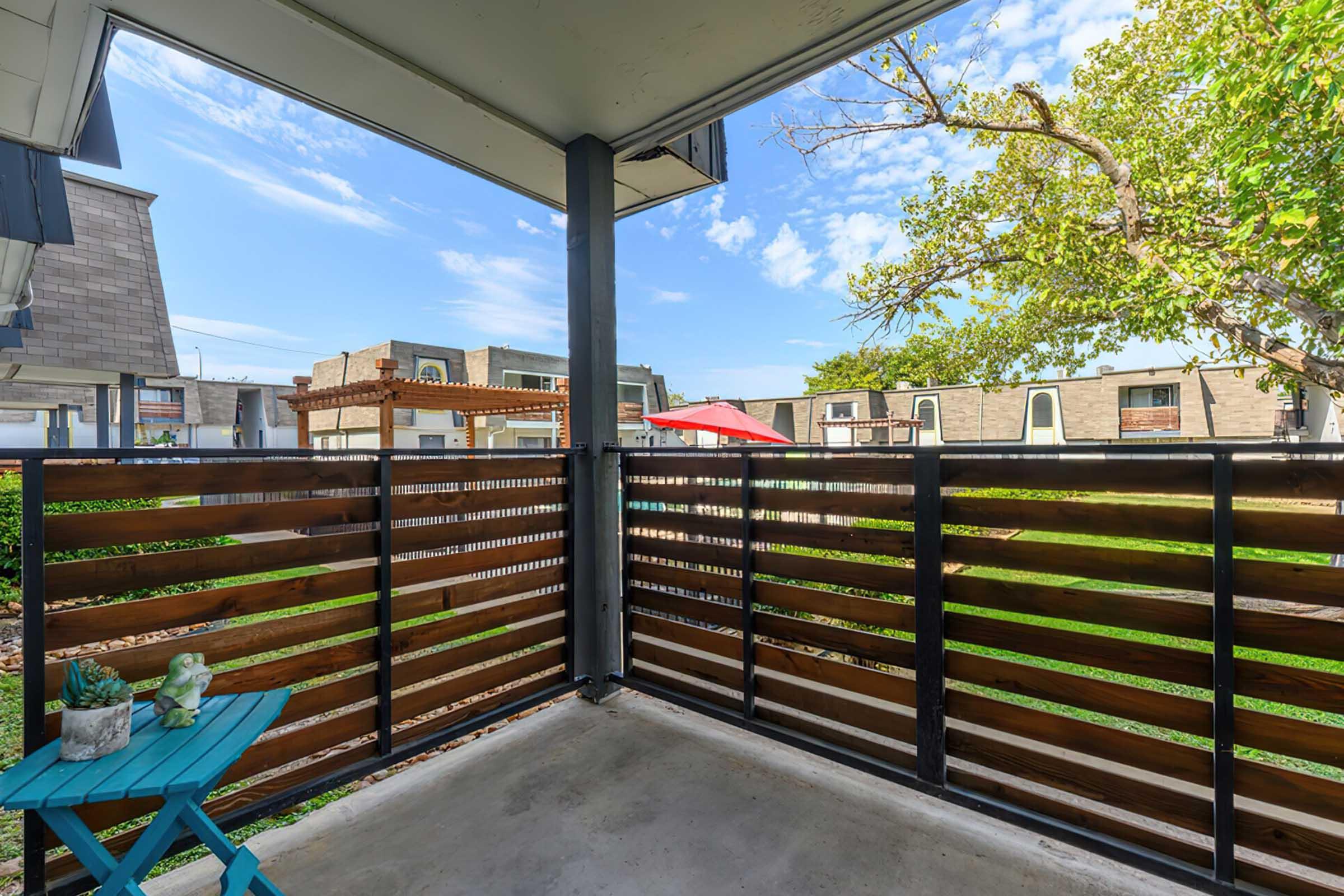 View from a balcony featuring a wooden railing, showcasing a sunny outdoor space with green grass and a tree. In the foreground, there is a small turquoise table with decorative items. The background includes buildings and a patio area with an umbrella, under a clear blue sky.