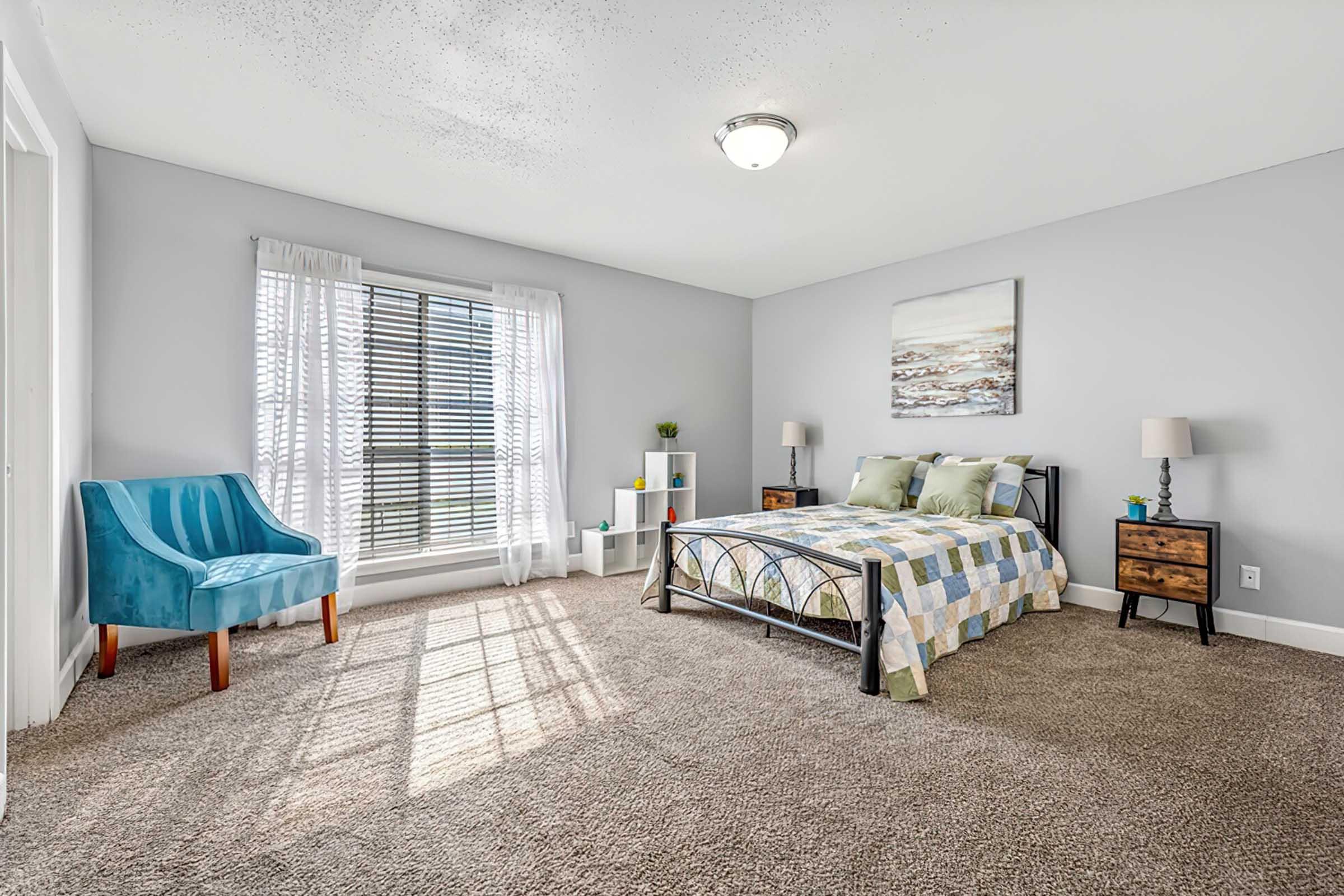 A bright and airy bedroom featuring a bed with a green and yellow patterned quilt, two bedside tables with lamps, a small bookshelf, and a blue chair. Natural light streams in through large windows with sheer curtains, highlighting the plush carpet and decorative wall art.