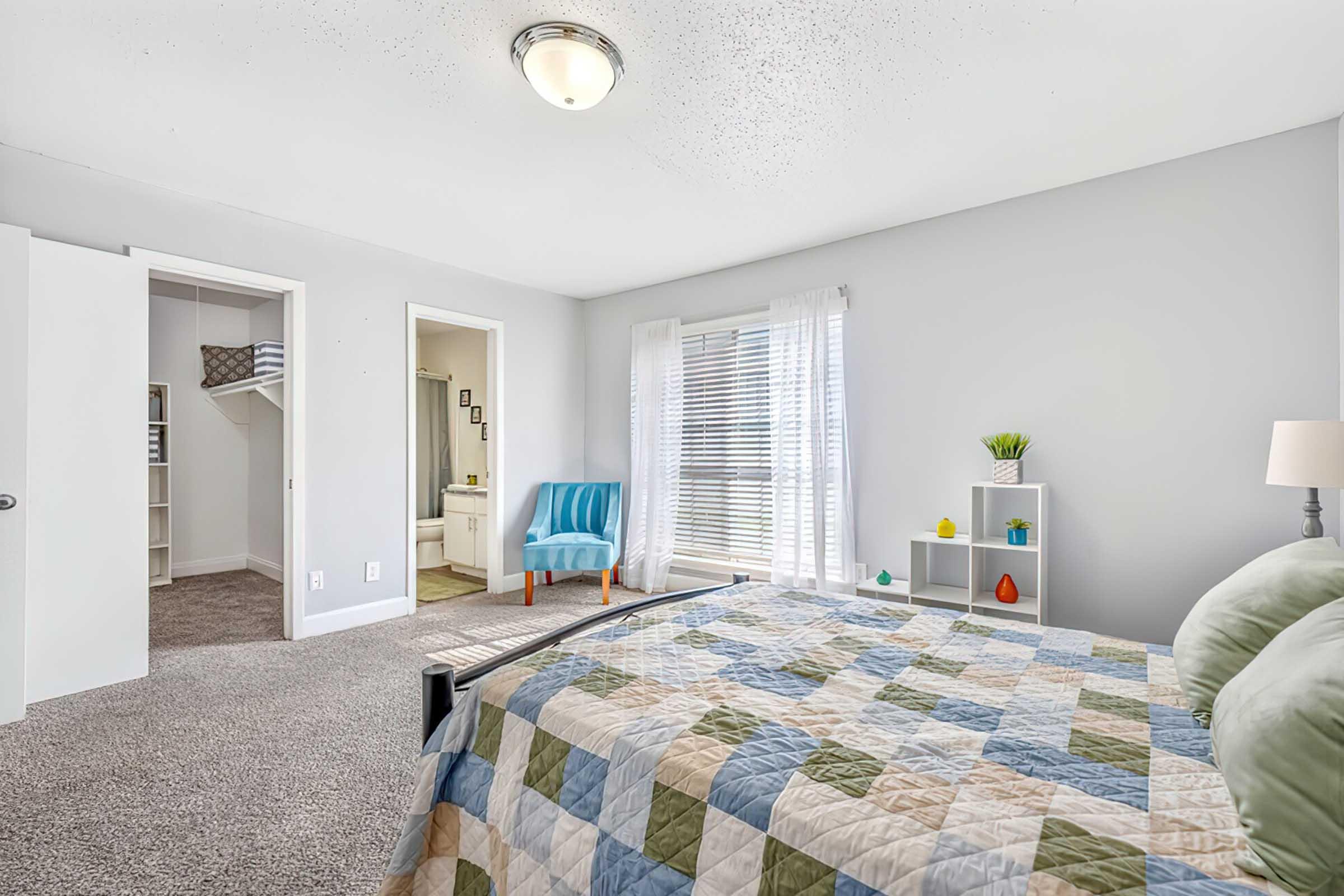 A bright and airy bedroom featuring a neatly made bed with a colorful quilt. A cozy blue chair is positioned near a window adorned with sheer curtains, while a small bookshelf with decorative items is nearby. A doorway leads to a closet and a bathroom. The room has light gray walls and soft carpet.
