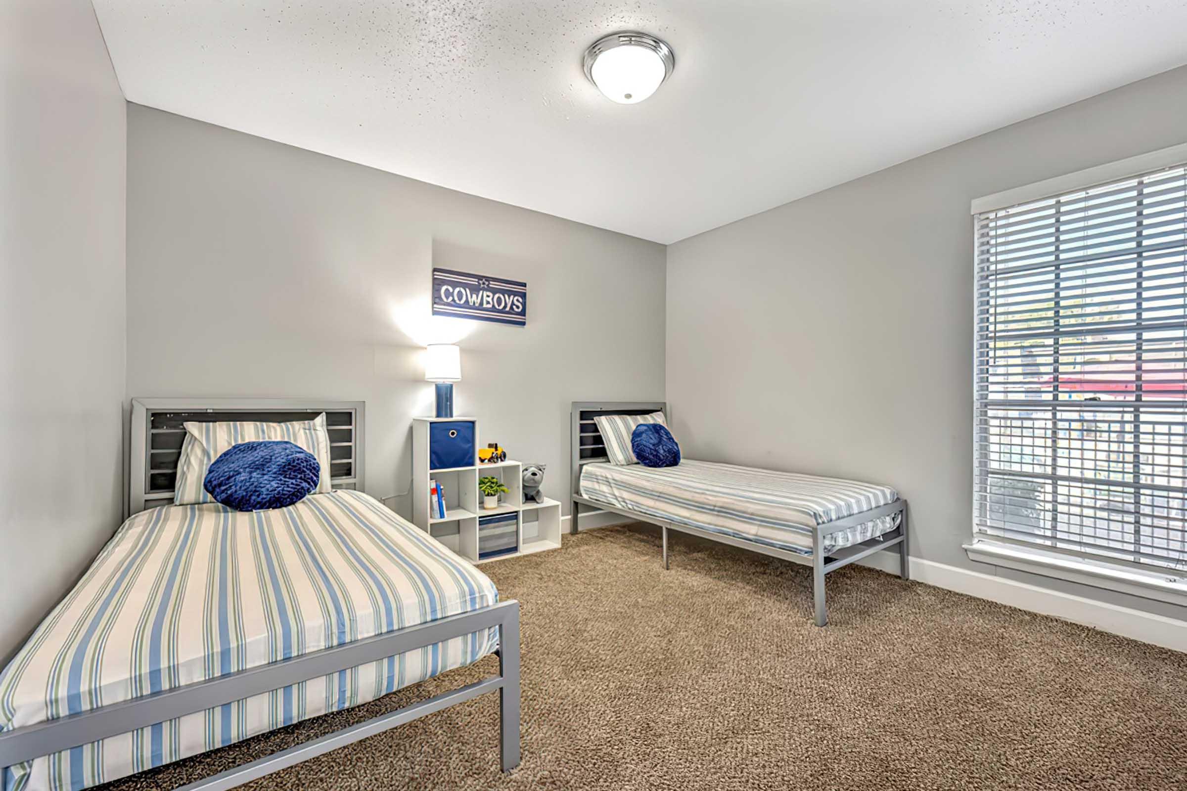 A cozy bedroom featuring two single beds with striped bedding, a nightstand with a lamp, and a decorative wall piece that says "COWBOYS." The room has light gray walls and a window with blinds that allow natural light, while carpet flooring adds warmth to the space.