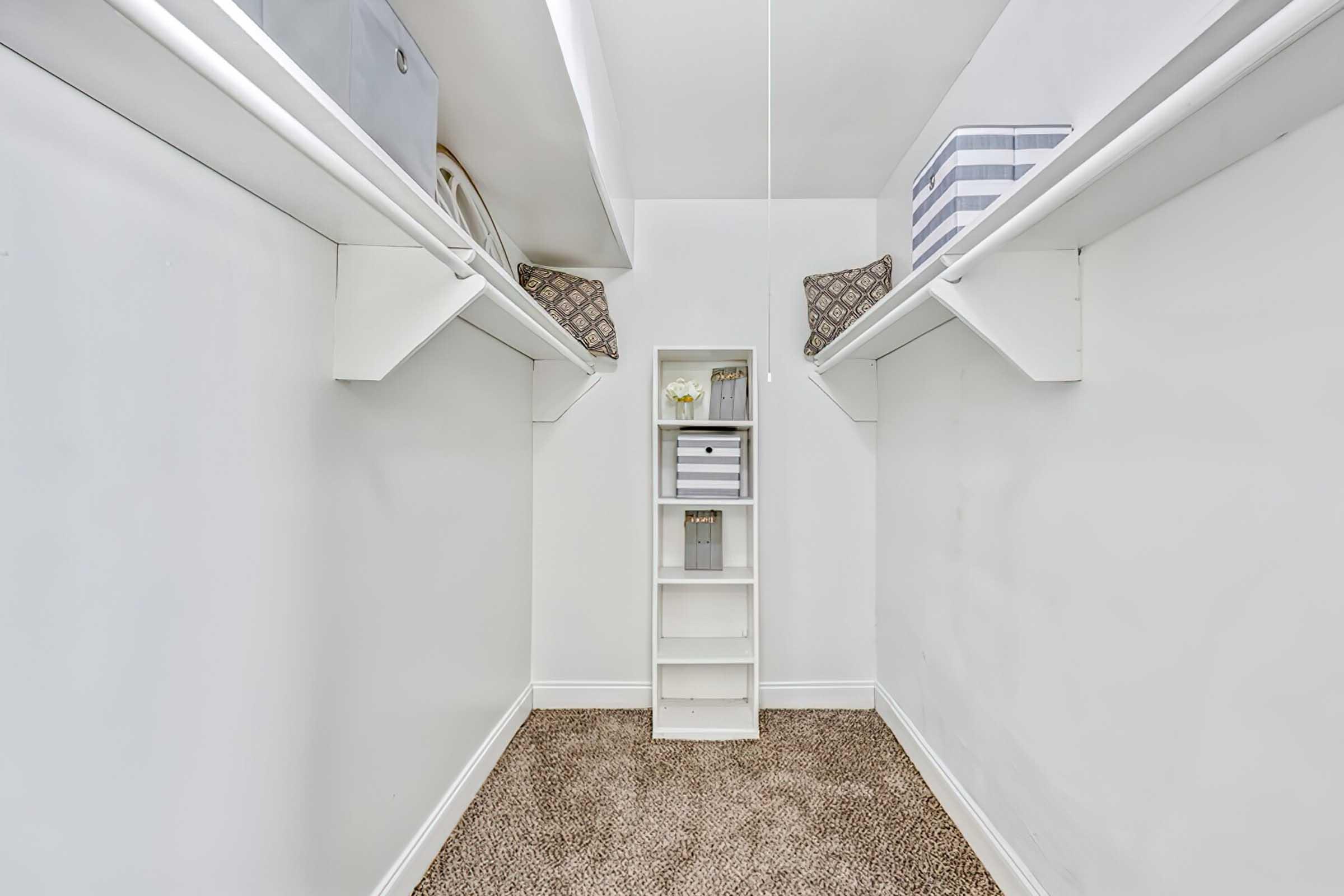 A view of a minimalist closet with white walls and two shelves on either side. The shelves hold decorative boxes, and a small white shelving unit with open compartments is centered at the back. The floor is covered with brown carpet, creating a clean and organized storage space.