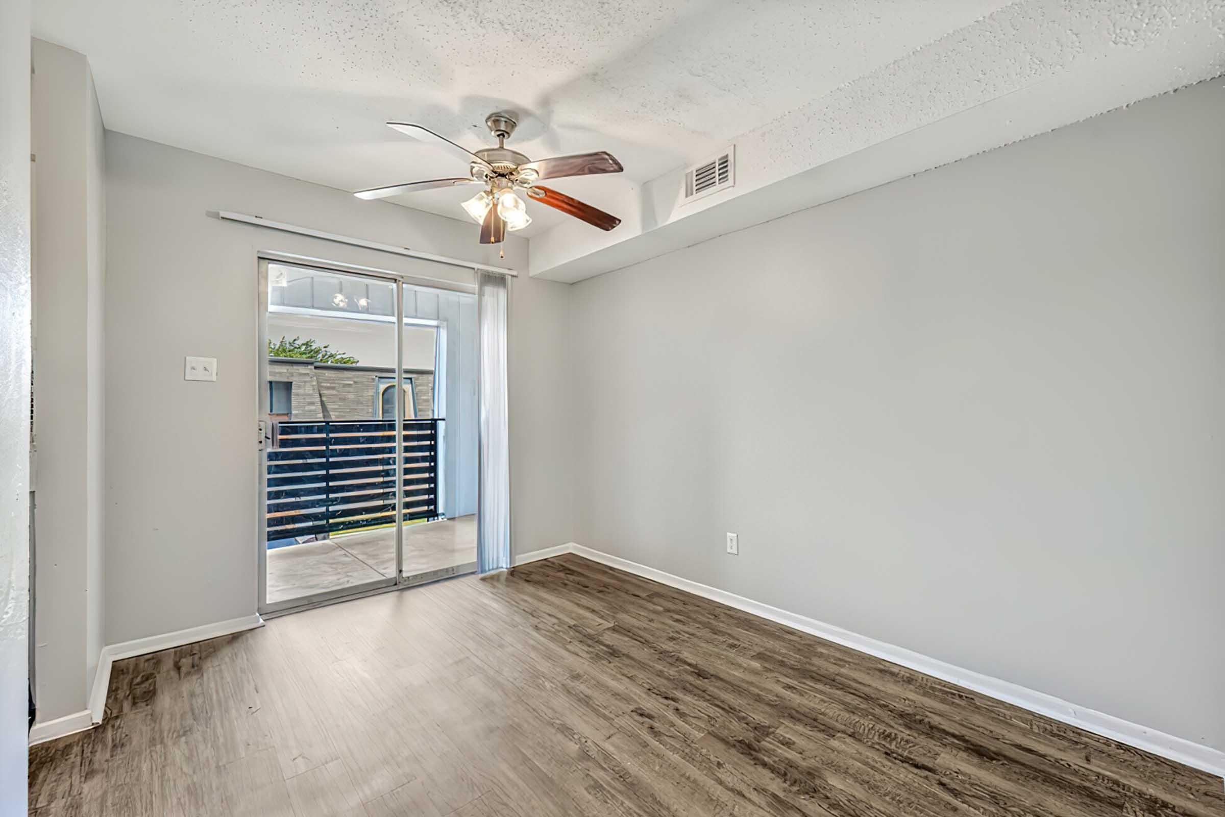 Interior view of a spacious room featuring a ceiling fan, light gray walls, and wood-like flooring. A glass sliding door leads to a small balcony area, allowing natural light to enter. The room is unfurnished, providing an open space ideal for various arrangements.
