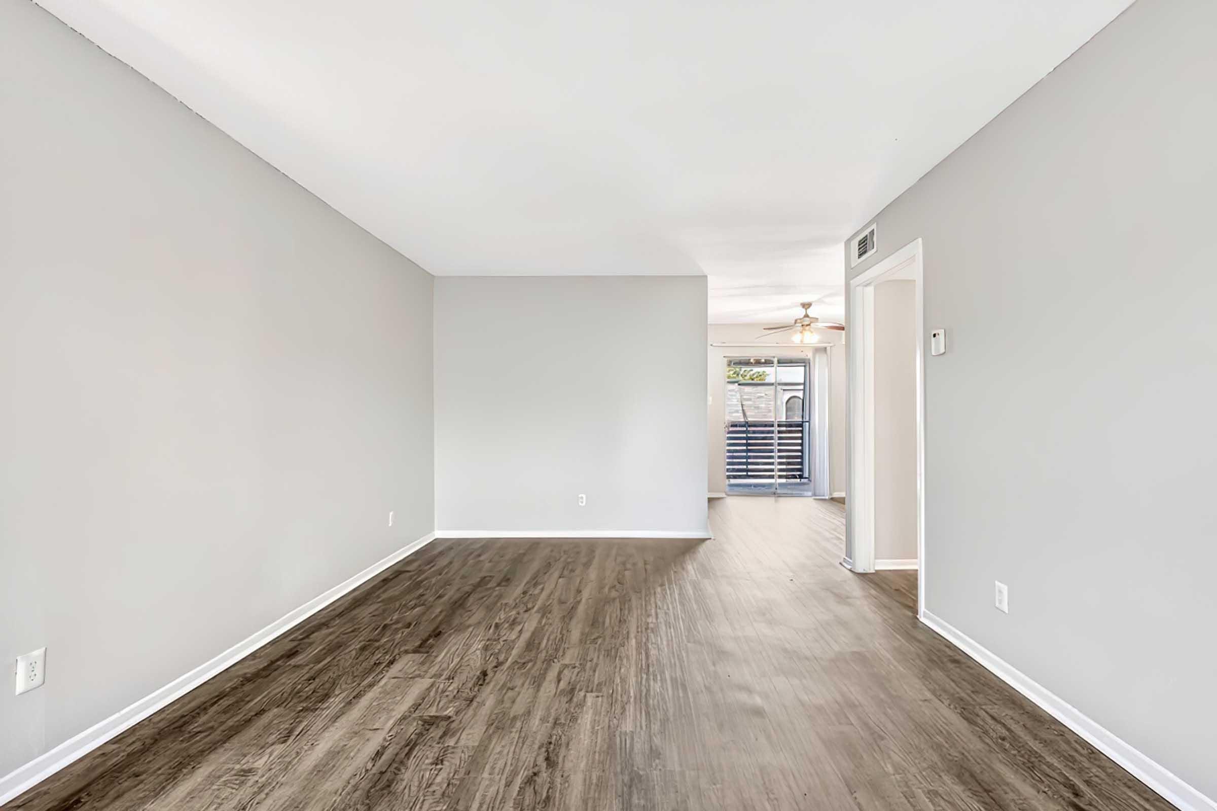 Empty interior of a modern apartment featuring gray walls, a wooden floor, and natural light coming from a doorway at the end. A ceiling fan is visible in the distance, and the layout suggests an open living area leading to a sliding door.