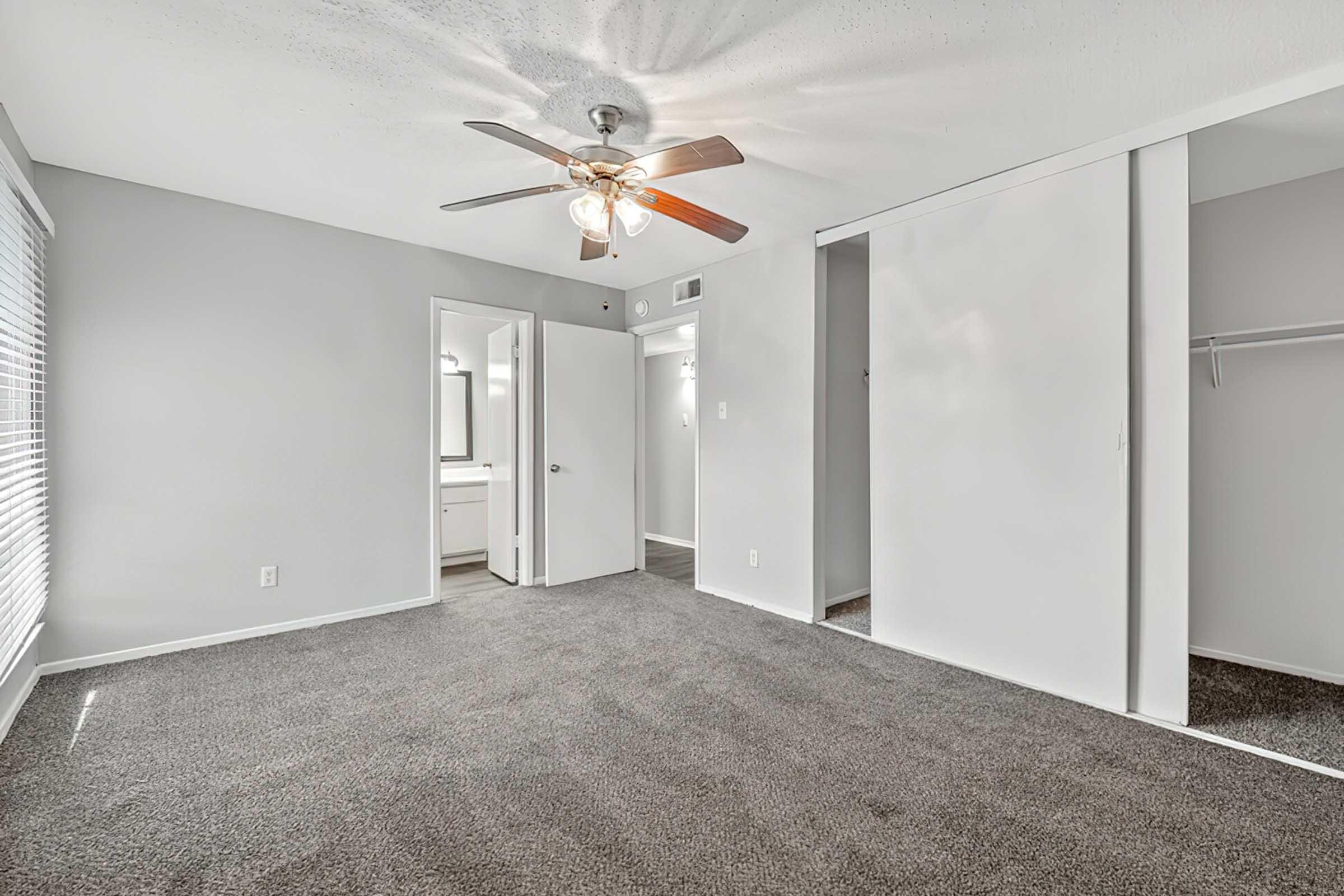 Spacious bedroom with gray carpeting, ceiling fan, and light gray walls. Features a doorway leading to a bathroom area and a closet space. Natural light streams through a window adorned with white blinds. The overall atmosphere is bright and inviting, perfect for a comfortable living space.