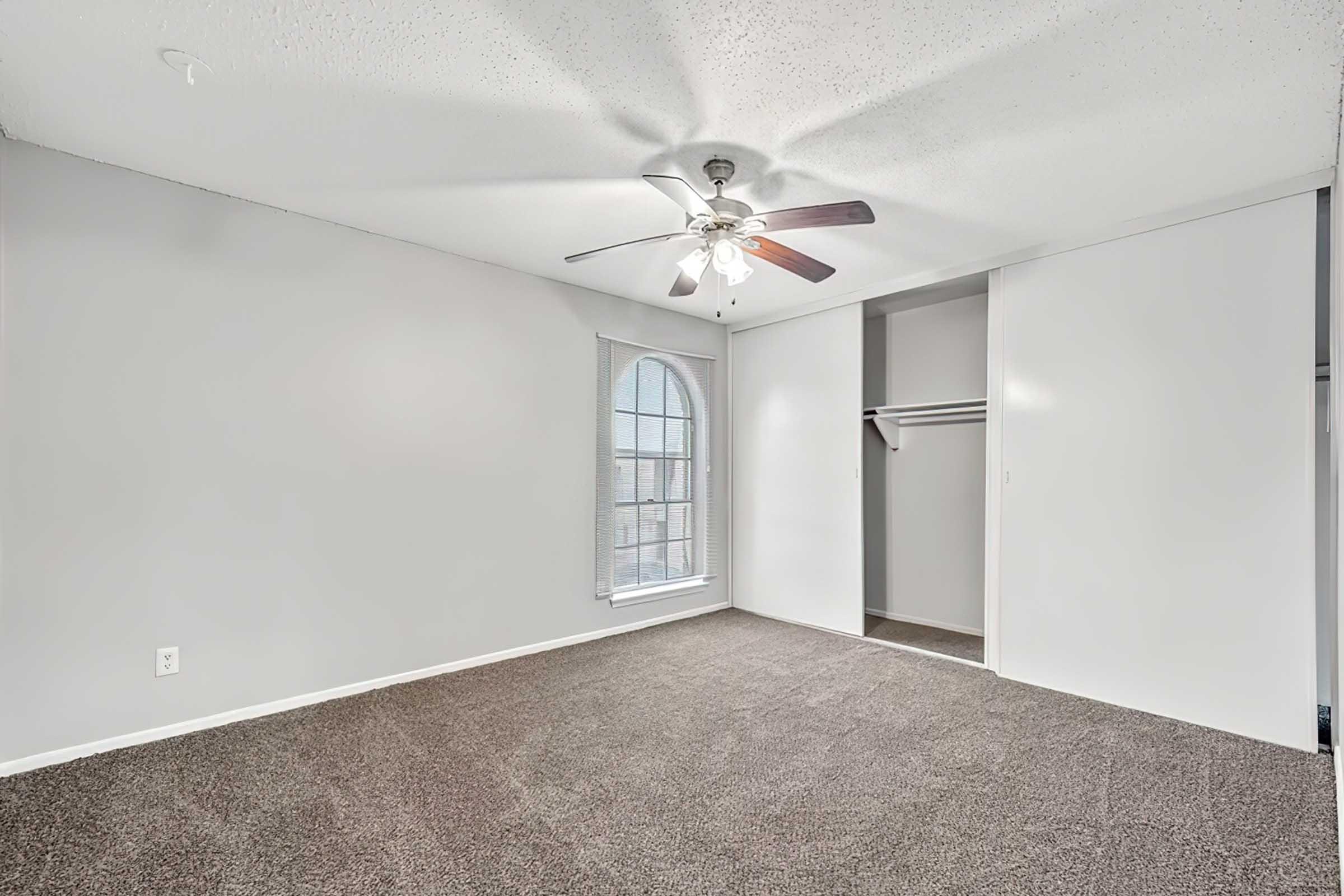Empty room with light gray walls and plush carpet. A ceiling fan is installed in the center, and there is a window with an arched design letting in natural light. On one side, a closet with sliding doors is visible, contributing to the spacious feel of the area.