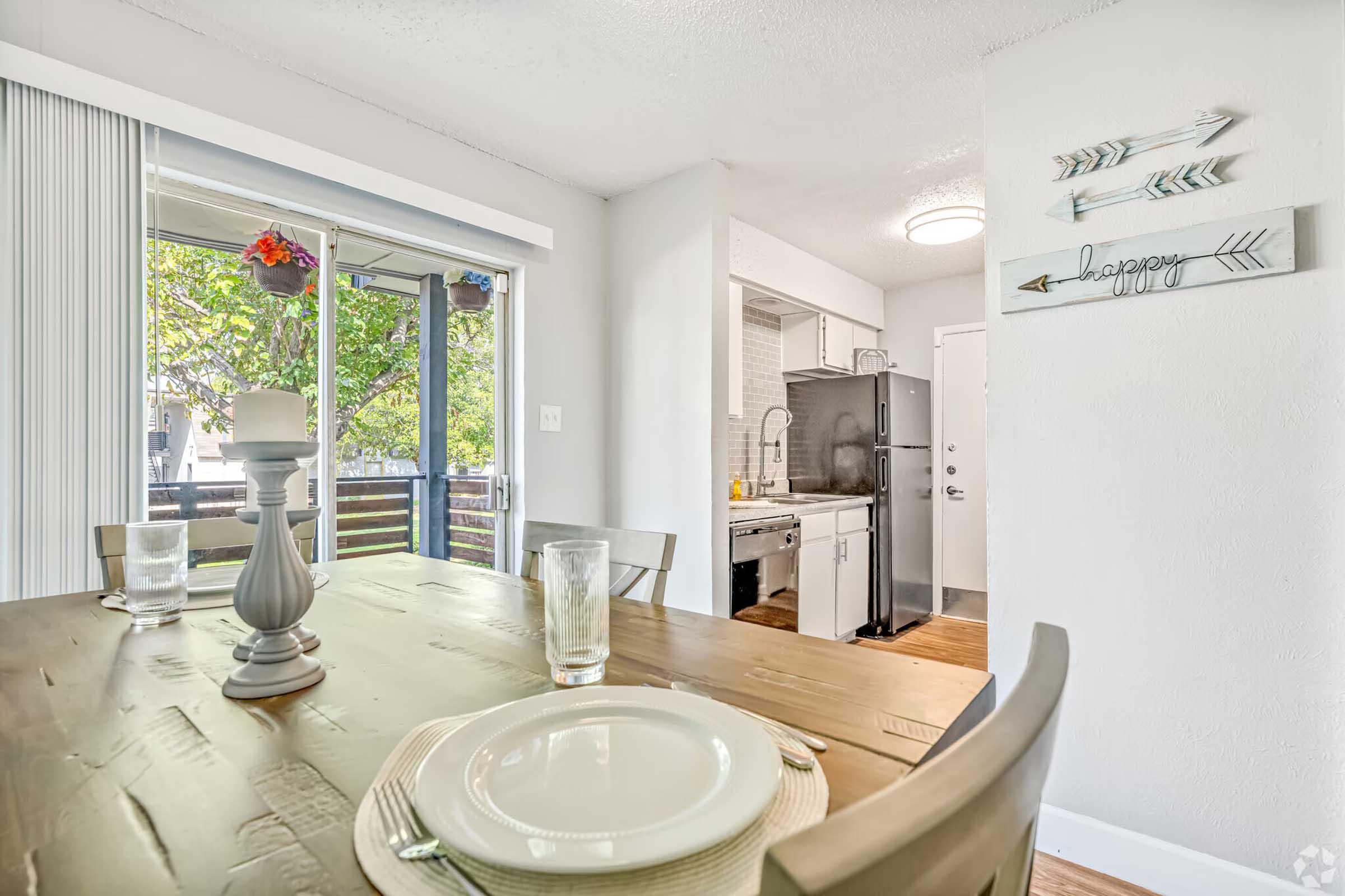 Cozy dining area with a wooden table set for a meal, featuring plates and glasses. In the background, a modern kitchen with stainless steel appliances is visible. Natural light streams in through a sliding glass door, revealing a balcony with greenery outside. Decorative arrows and a candle add warmth to the space.