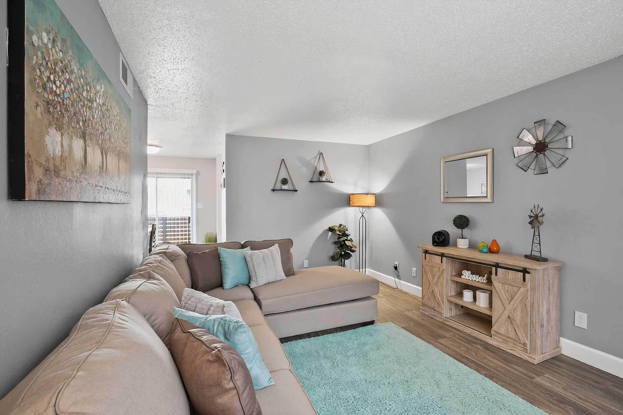 Cozy living room featuring a light gray wall, a large beige sectional sofa with accent pillows, and a decorative teal area rug. There is a wooden console table with decorative items, a mirror, and a wall clock. The space is adorned with wall art and geometric decorations, creating a welcoming and stylish atmosphere.