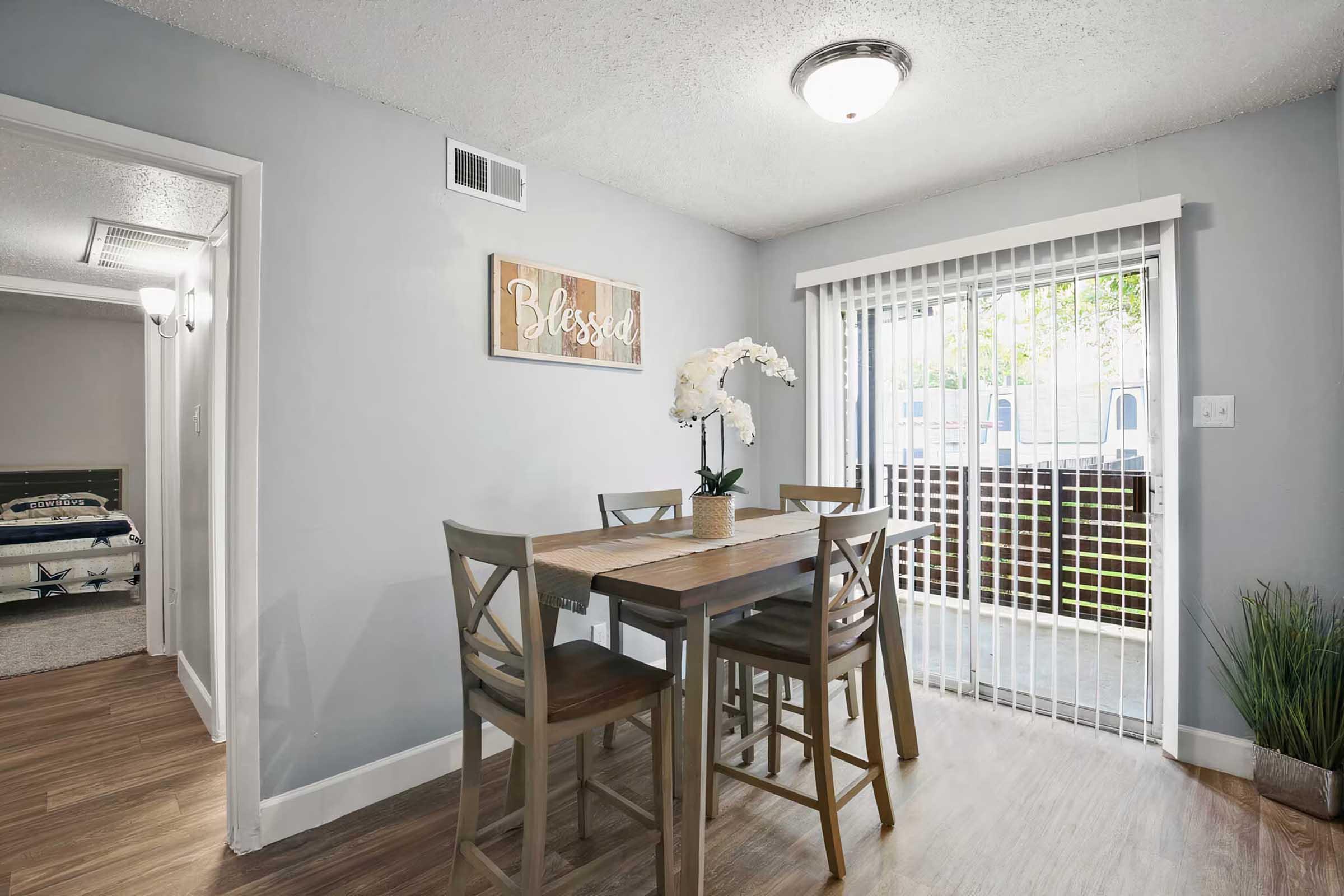 A cozy dining area with a wooden table and four chairs, adorned with a vase of white orchids. A decorative sign reading "Blessed" hangs on the wall. In the background, sliding glass doors lead to a balcony, and warm lighting enhances the inviting atmosphere. The floor features a stylish wood-like finish.