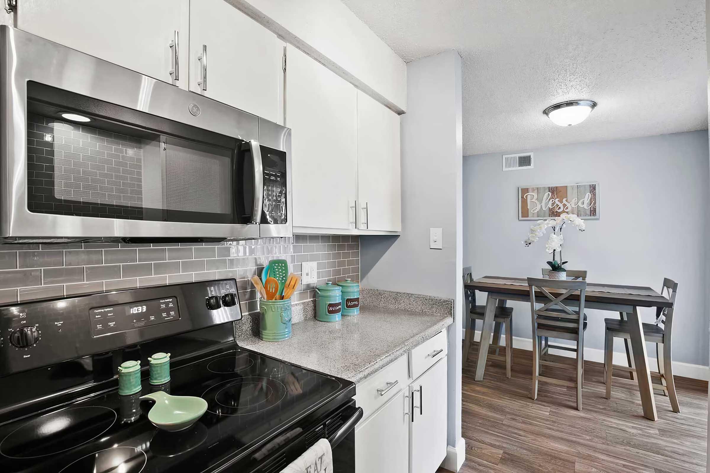 Modern kitchen featuring stainless steel appliances, including a microwave and stove. Grey tile backsplash complements light cabinets. In the background, a dining area with a wooden table and decorative flower centerpiece is visible, along with a wall sign that says "Blessed."