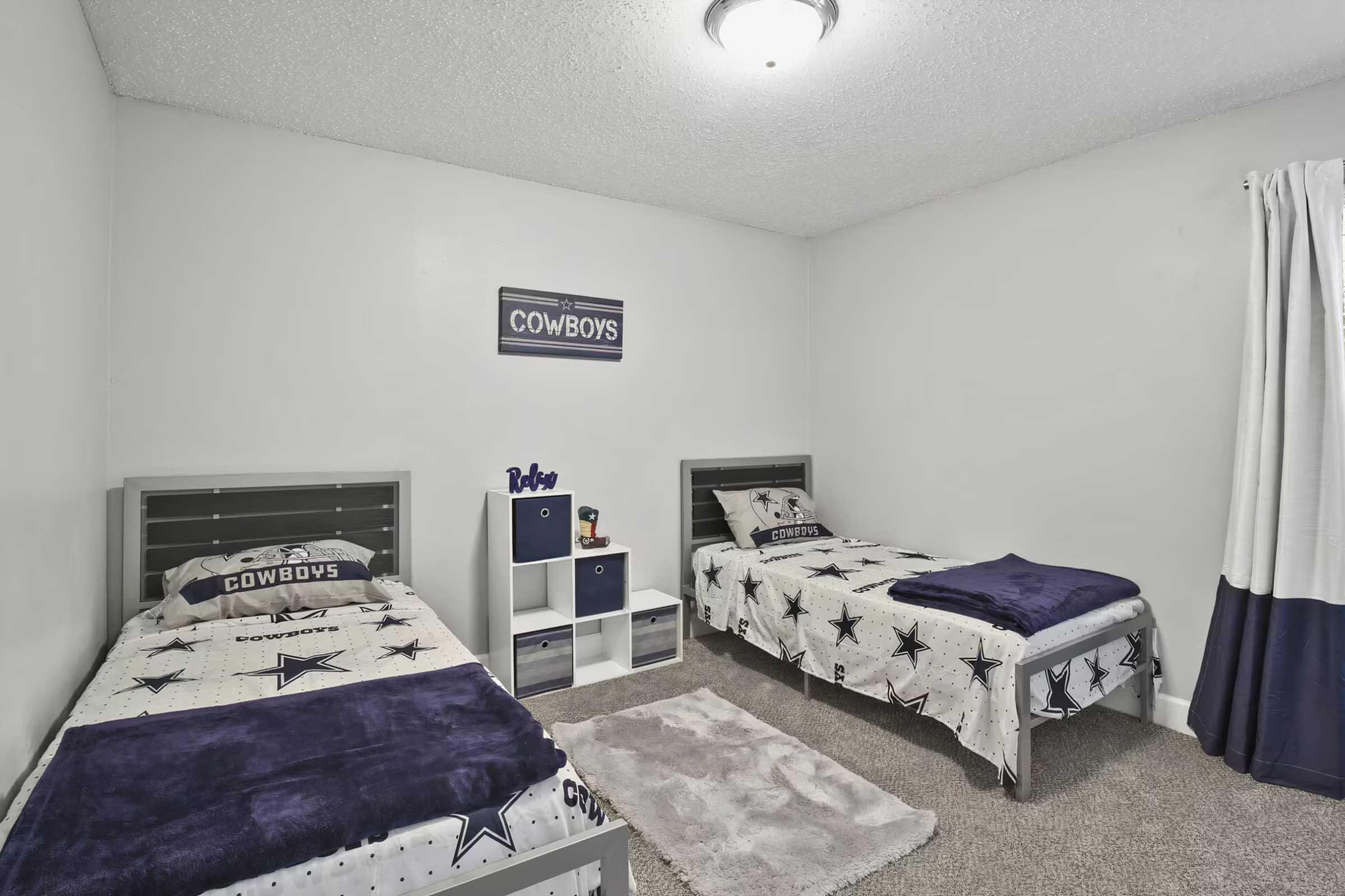 A cozy bedroom featuring two twin beds with Dallas Cowboys-themed bedding. The room has a neutral-colored wall, a small shelf with decorative items, and a soft rug on carpeted flooring. Natural light enters through a window with a curtain, creating a welcoming atmosphere.