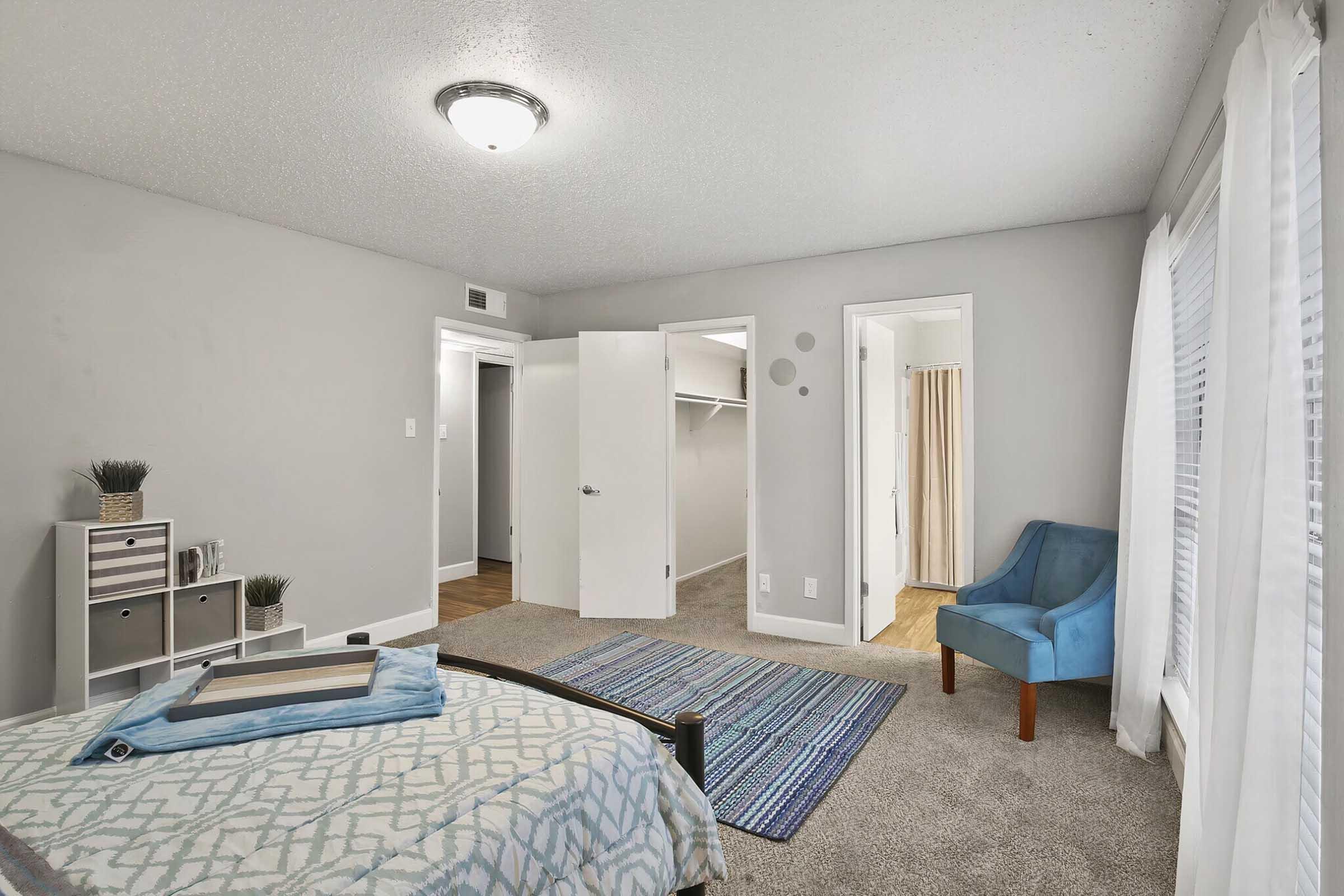 A cozy bedroom featuring a bed with a patterned duvet, a blue accent chair, and a small bookshelf. The walls are painted in a light gray color, and there is a large window with white curtains allowing natural light in. A closet and a bathroom doorway are visible in the background. Soft carpet flooring completes the inviting atmosphere.